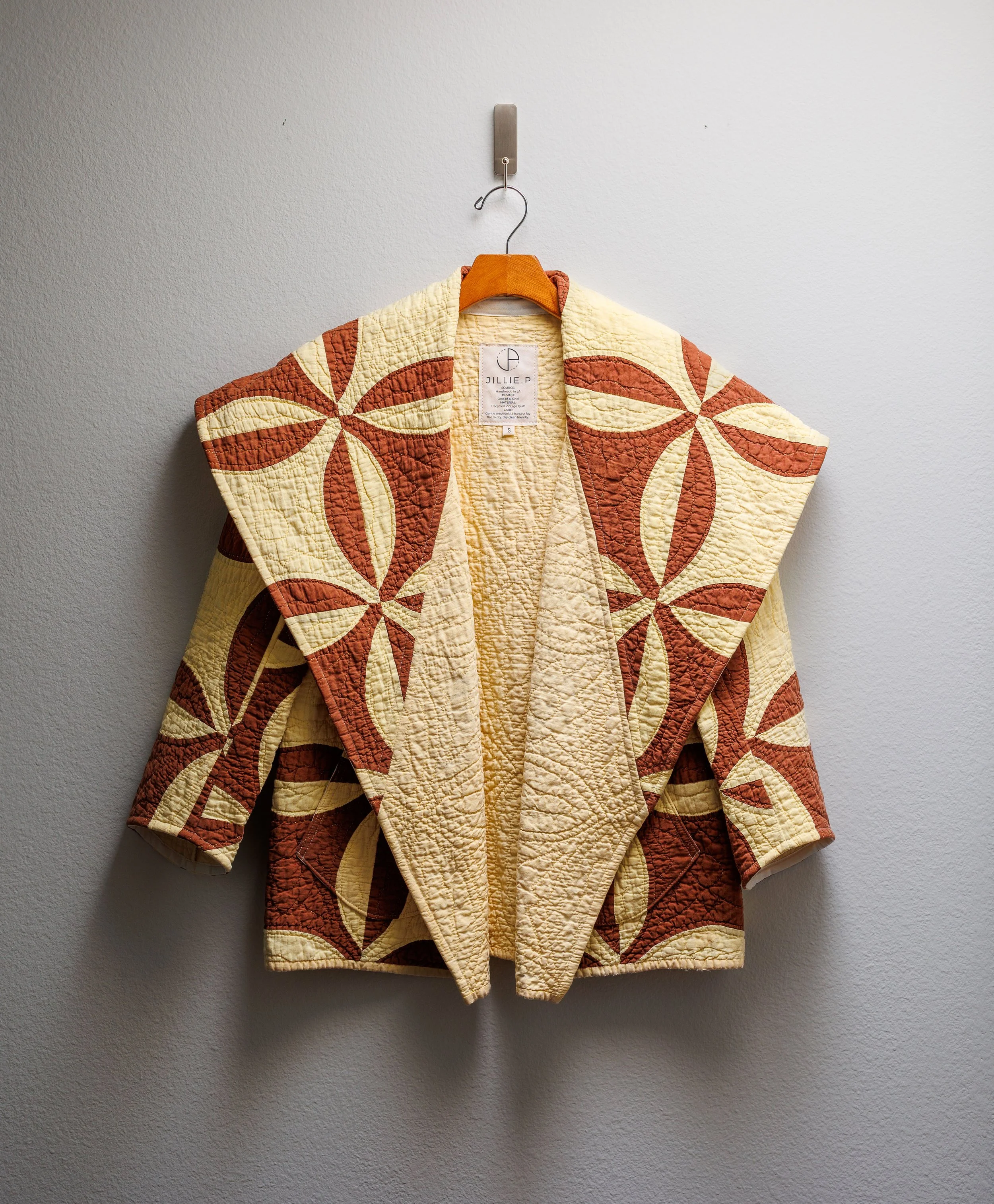 A quilted jacket with a floral pattern in warm colors hanging on a wooden hanger against a plain white wall.