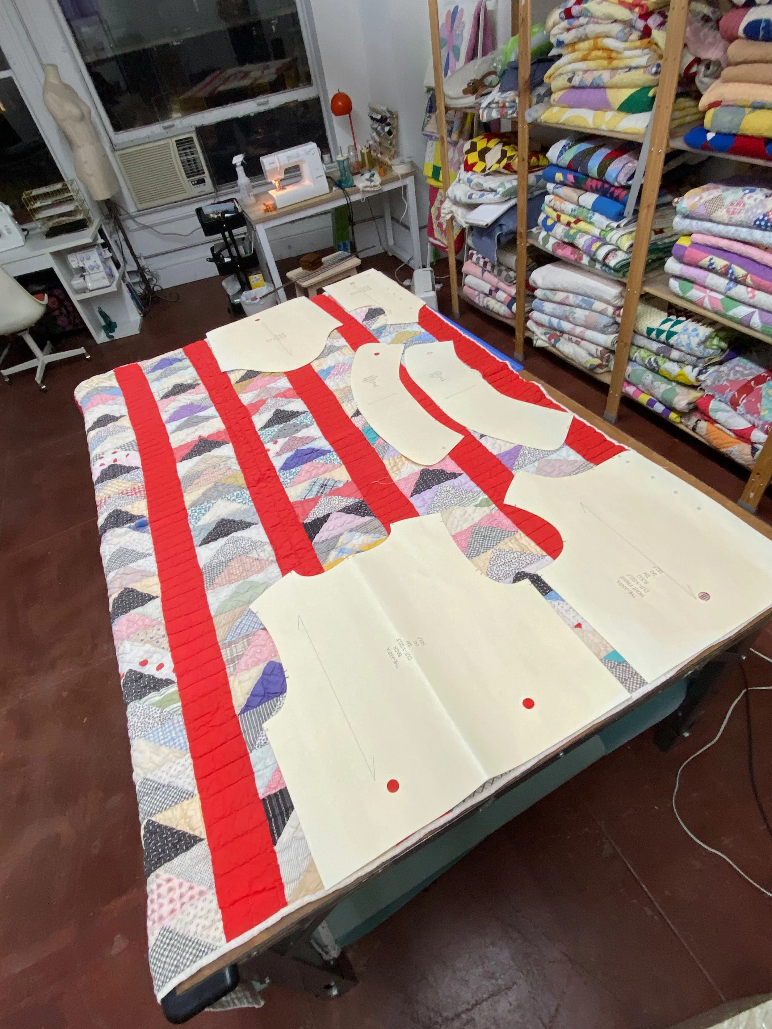 A sewing workspace with a quilt laid on the table, sewing patterns and fabric, along with shelves holding stacks of folded fabric and quilts, and a corner with sewing equipment.