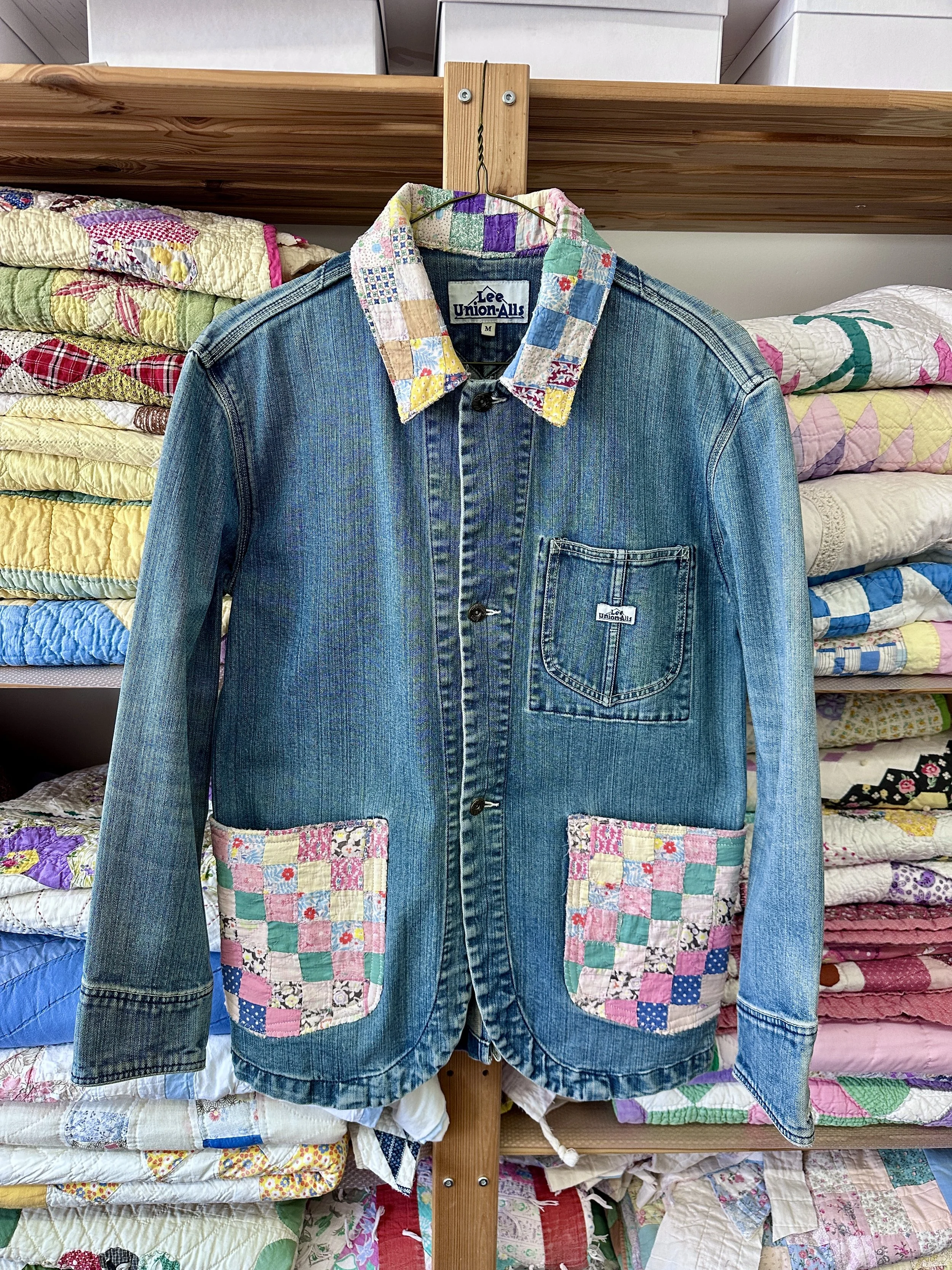 A denim jacket with patchwork quilt pockets hanging on a wooden rack, surrounded by folded quilts on shelves.