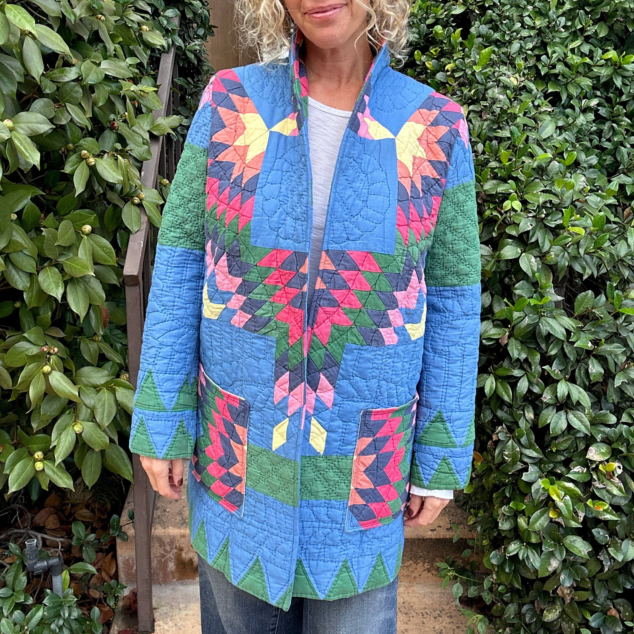 A woman wearing a colorful quilted jacket with a geometric pattern of pink, yellow, blue, green, and purple standing outdoors. The background includes green bushes and a wooden fence.