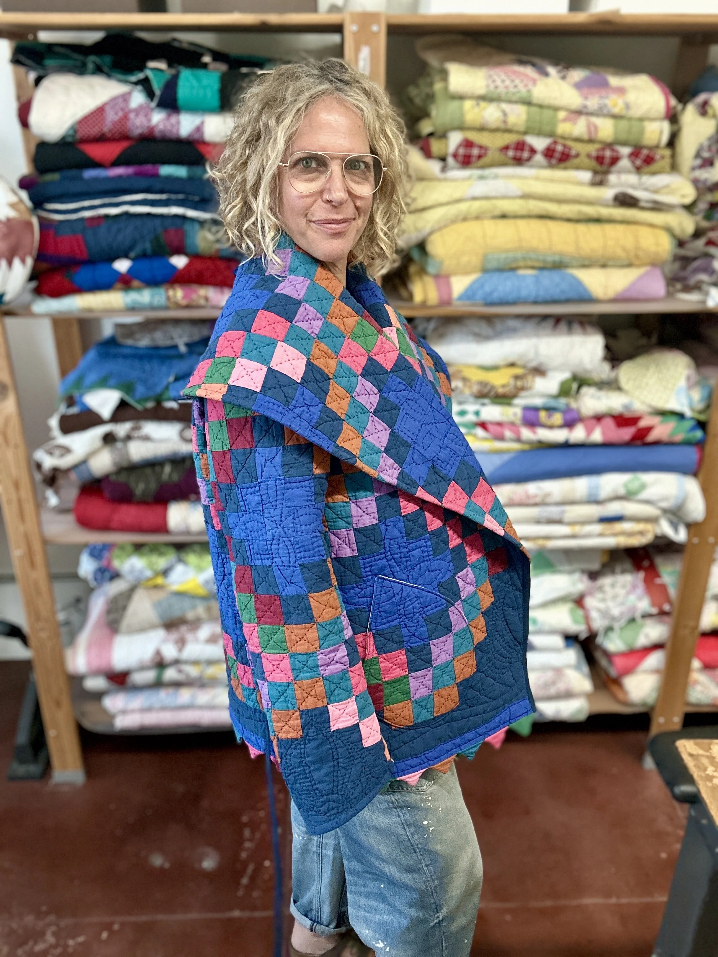 Person with curly blonde hair wearing glasses and a colorful patchwork jacket standing in front of stacks of folded quilts on shelves.