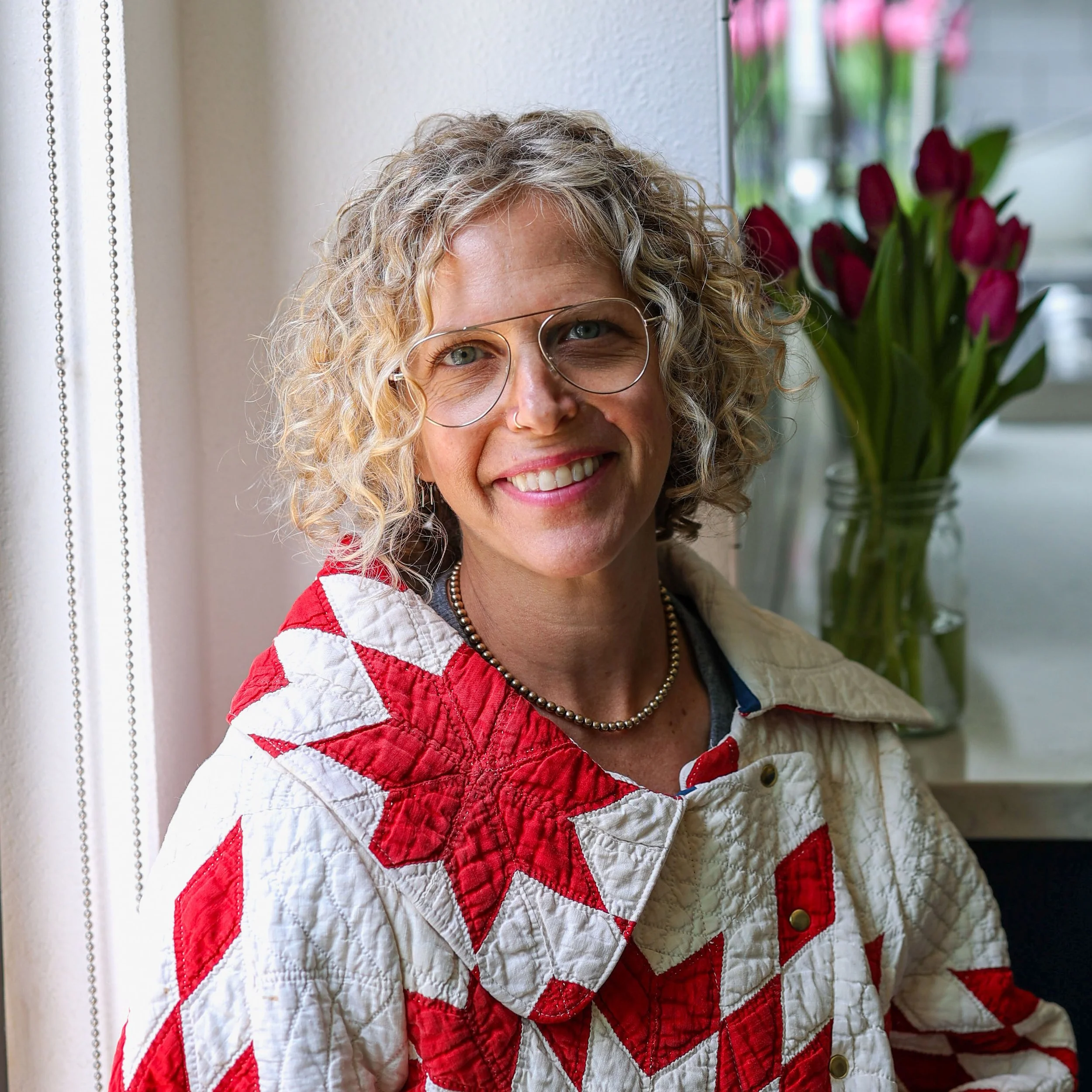 Close-up of a smiling woman with curly blonde hair wearing glasses, a red and white quilted jacket, and a pearl necklace, standing next to a window with pink flowers in the background.