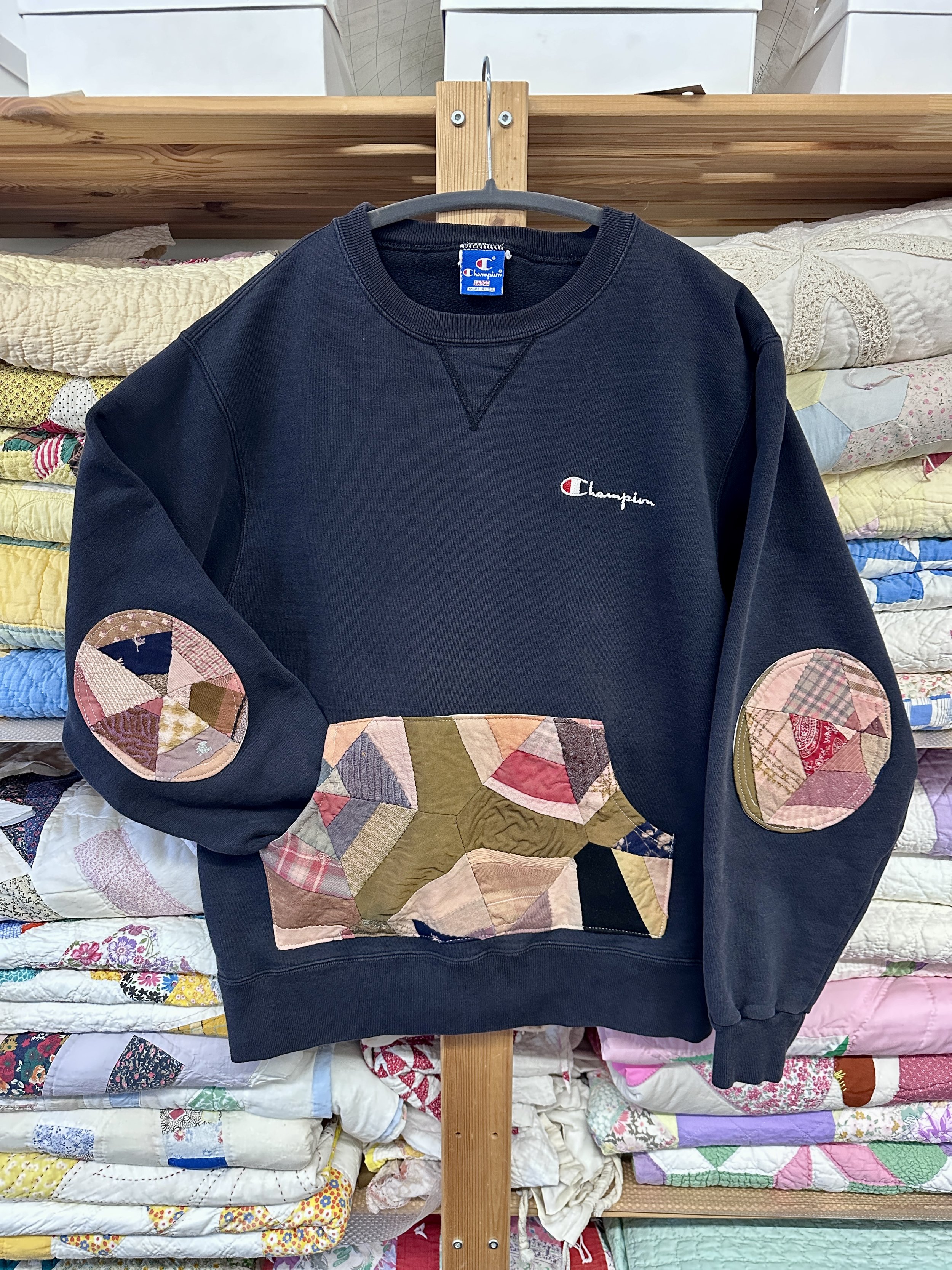 A blue Champion sweatshirt with quilted patches on the elbows hanging on a gray hanger in front of a stack of various quilts.