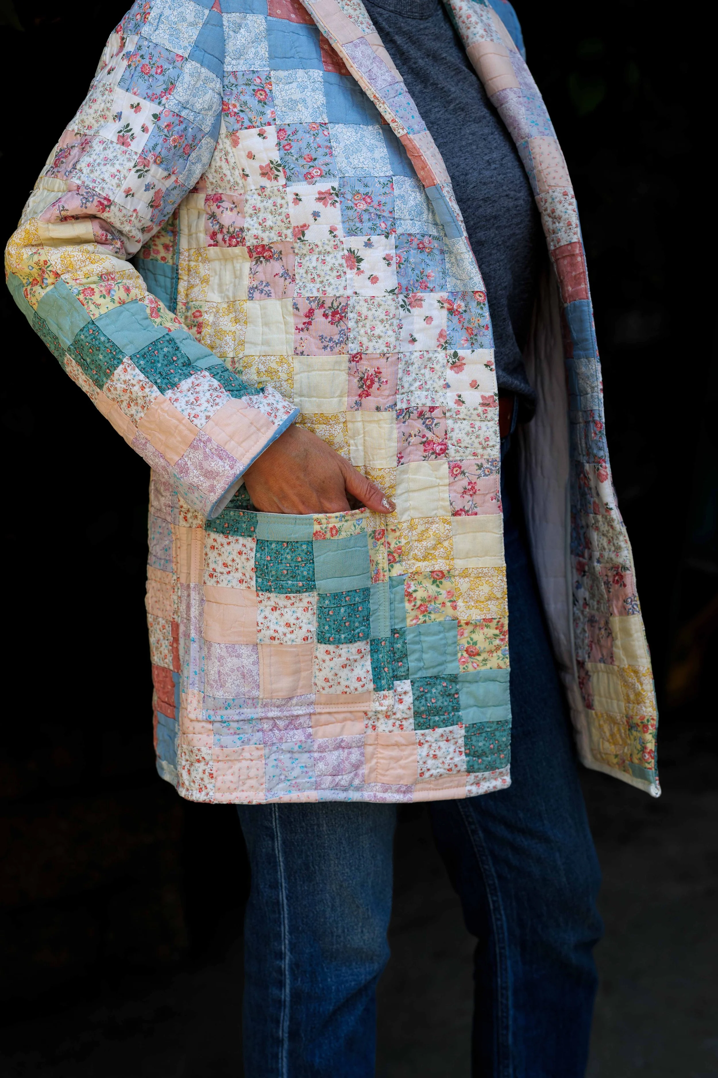 Person wearing a pastel-colored patchwork quilted coat over a dark gray shirt and blue jeans.