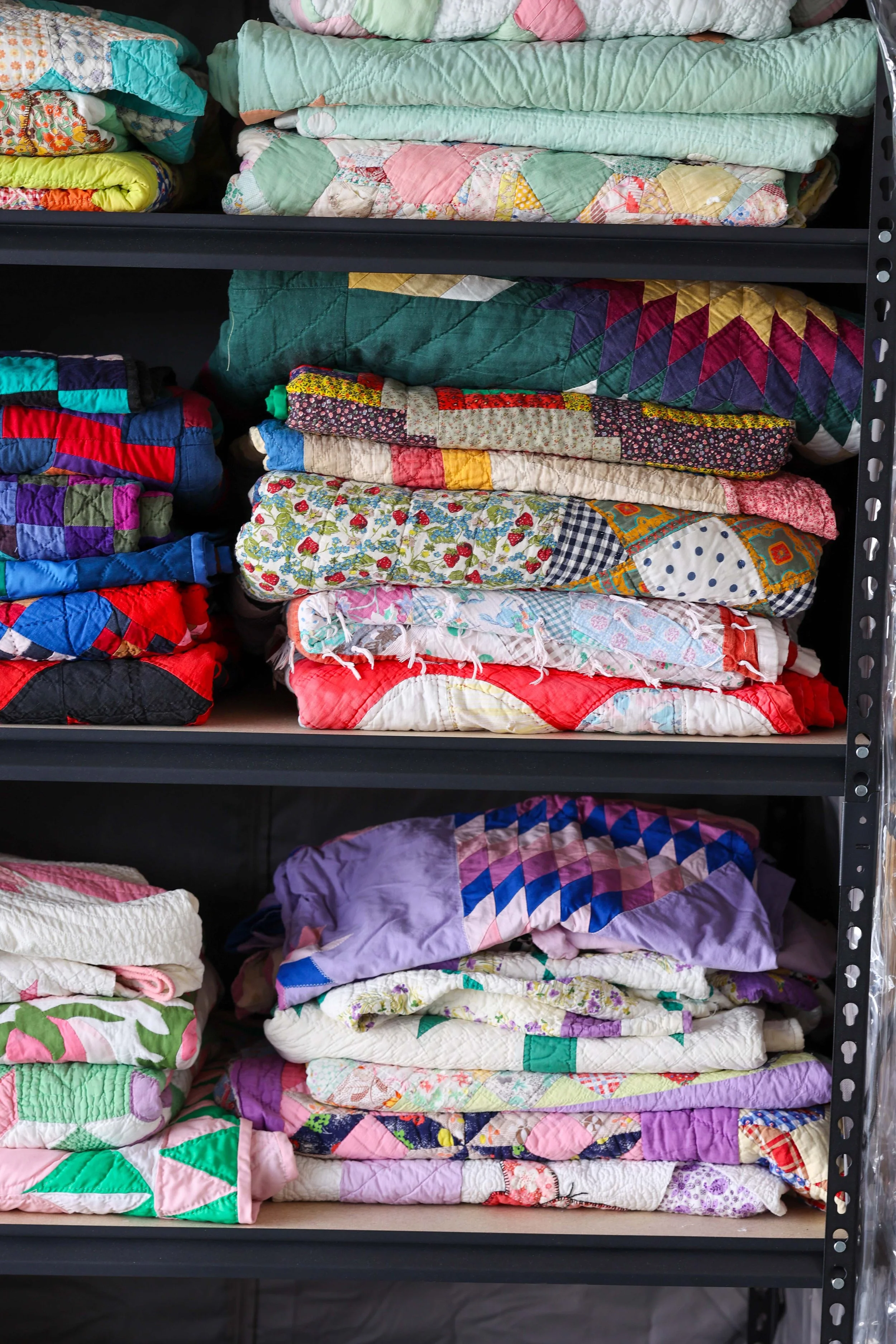 Stacked colorful quilts on metal shelves, various patterns including floral, geometric, and patchwork.
