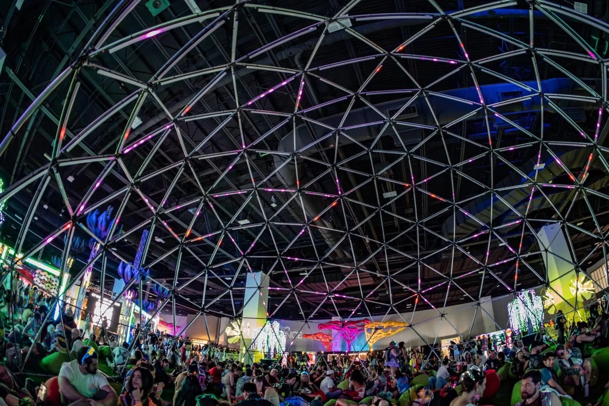 Inside a large, modern indoor festival or event space with a geodesic dome ceiling, colorful LED lights, and many people seated and walking around.
