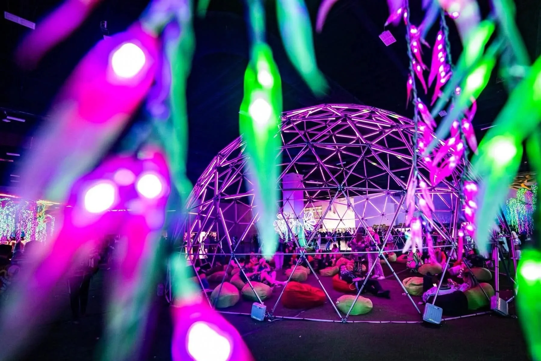 Colorful illuminated geodesic dome structure at a nighttime outdoor event with people seated underneath, seen through neon leaves in the foreground.