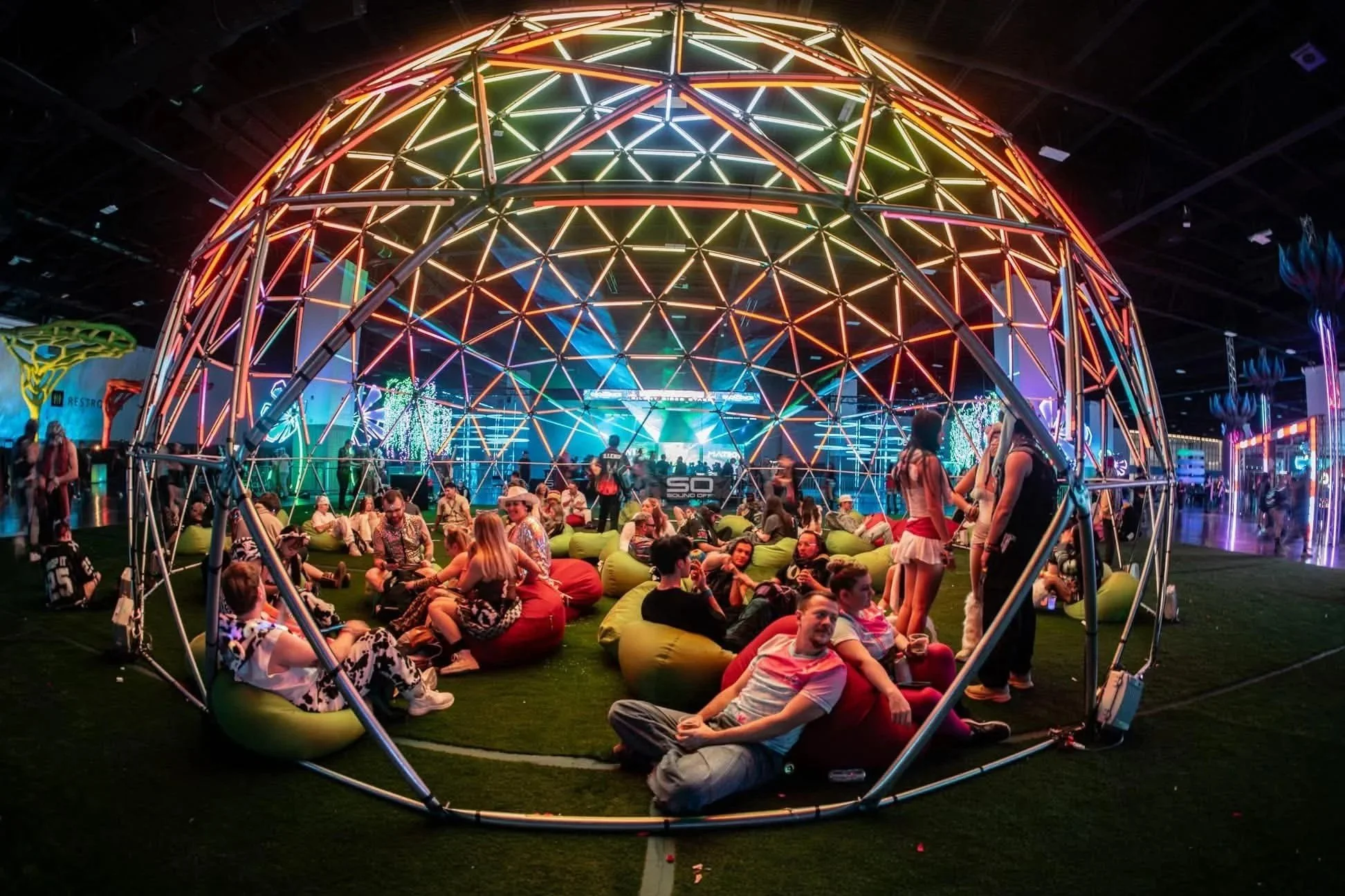 Colorful geometric dome structure with people sitting on bean bags inside at a lively festival with bright lights and a stage in the background.