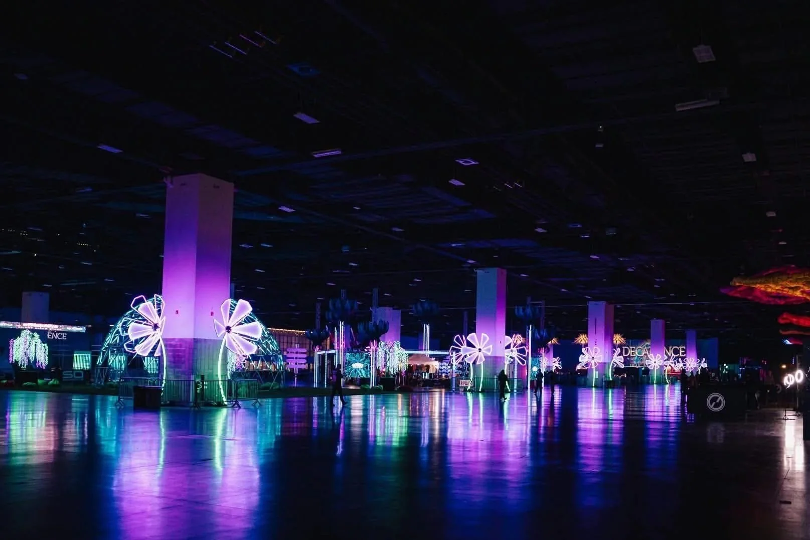 A dark indoor space with bright, colorful neon lights and decorations resembling large flowers. There are several people walking around in the illuminated area, with reflections on the shiny floor.