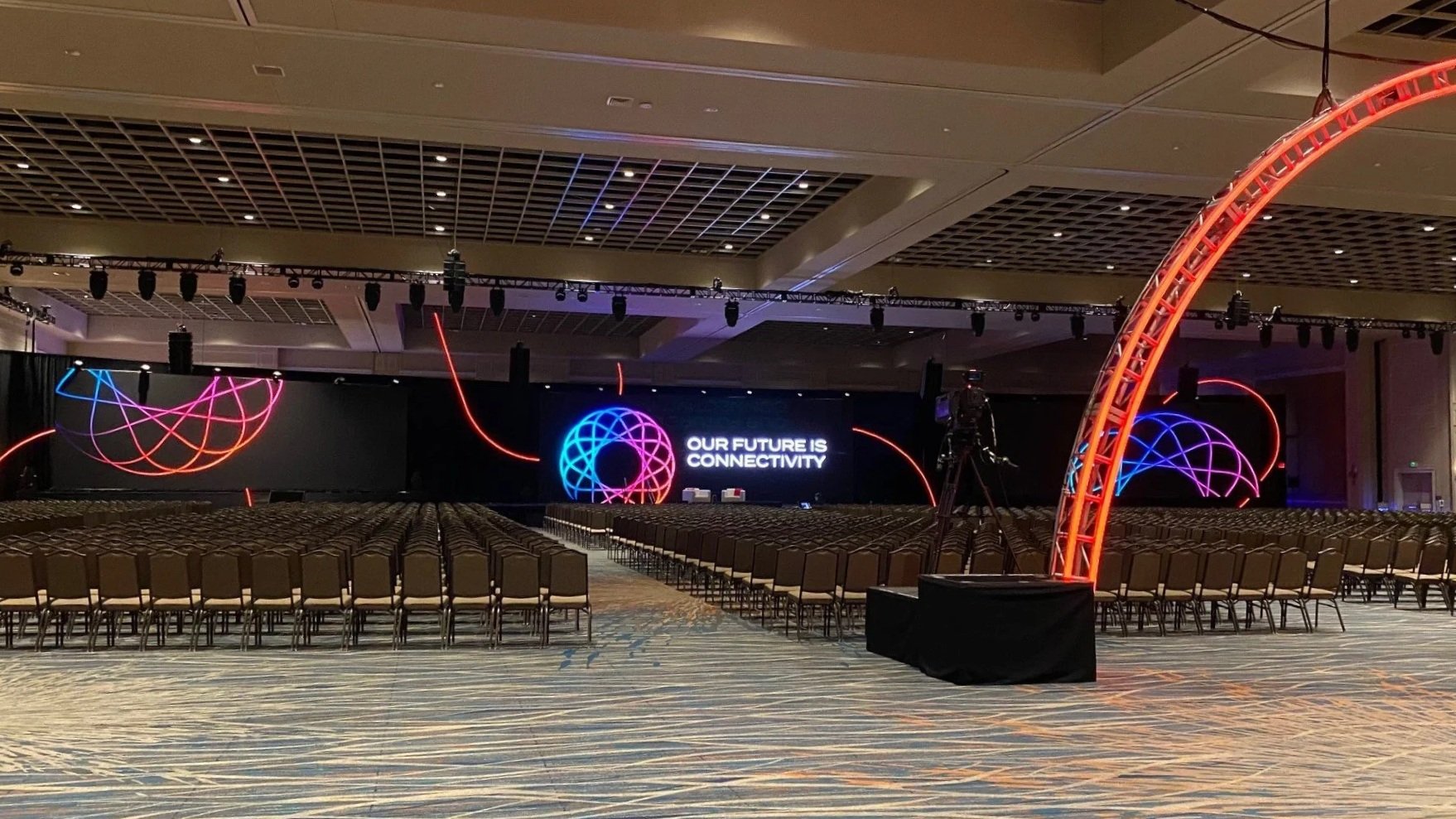 Conference room setup with rows of chairs facing a large stage featuring colorful LED lights and a screen displaying the message "Our Future is Connectivity.", Electronic Sales and Production, Denver, CO.