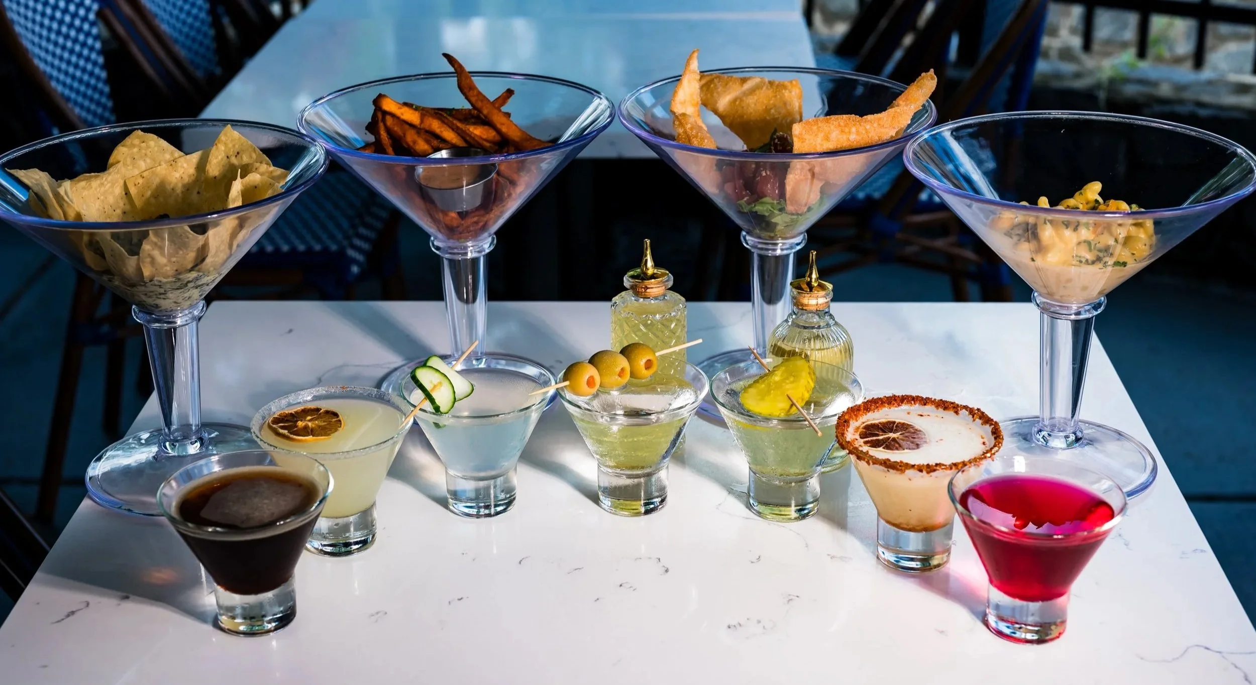 White marble table displaying four large martini glasses filled with appetizers and eight smaller garnished cocktails, set in an outdoor dining area with blue and white chairs.