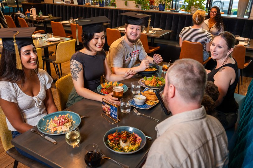 Group of diners wearing graduation caps seated around a restaurant table, eating plated meals and drinks in a modern indoor dining space.