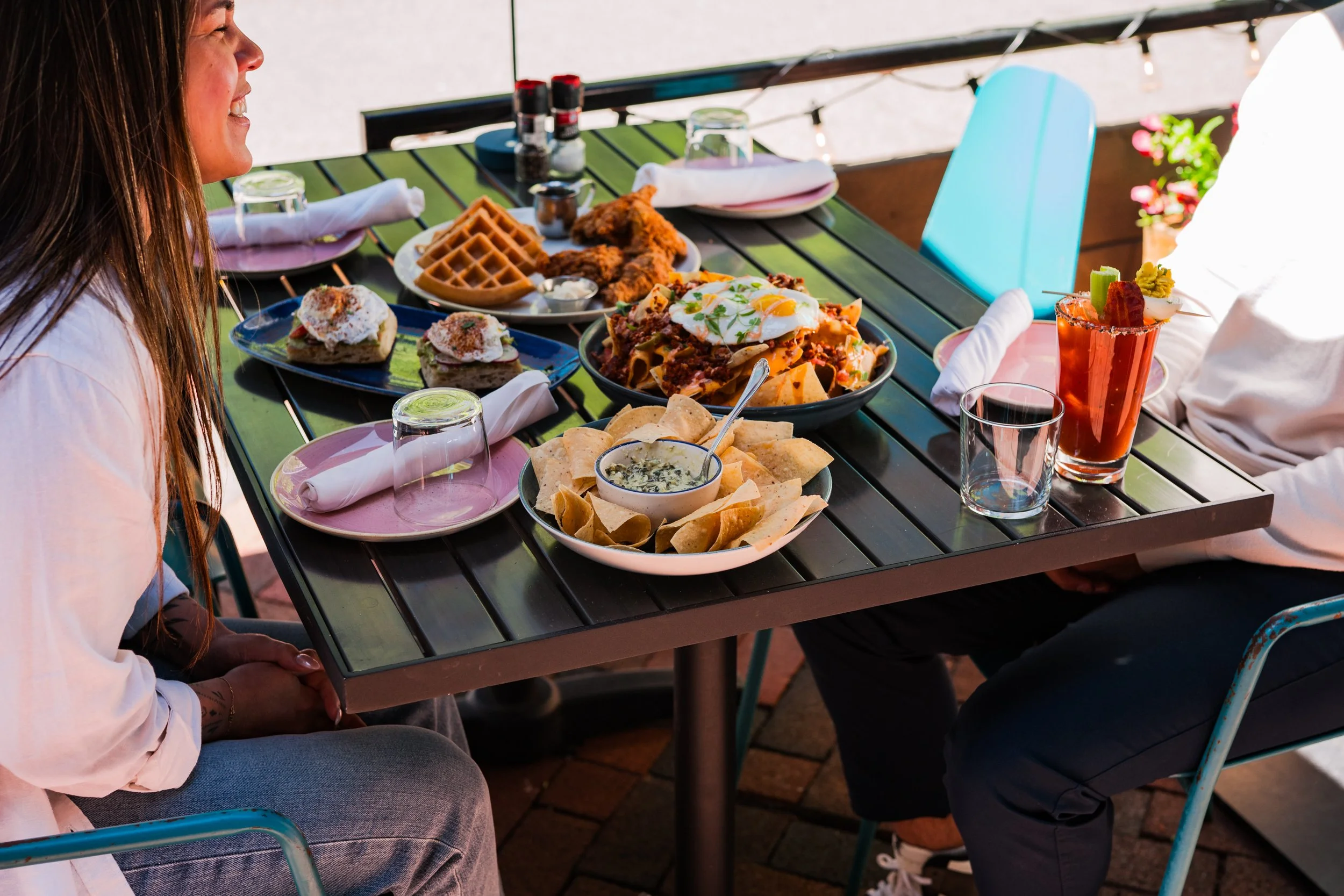 Outdoor dining table with shared brunch dishes including waffles, fried chicken, chips and dip, and cocktails as two people sit and talk.