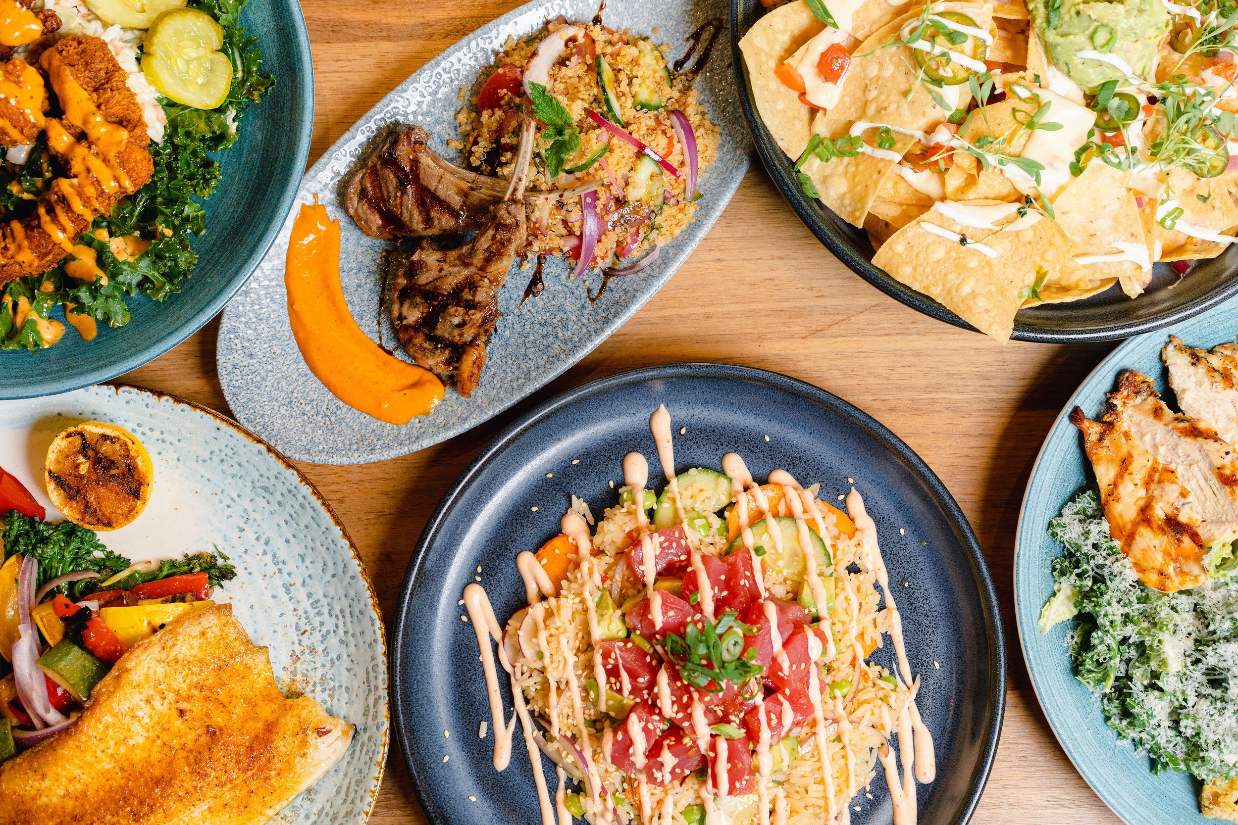 Assorted dishes on a wooden table, including nachos, grilled meats, salads, and grain bowls with colorful toppings.