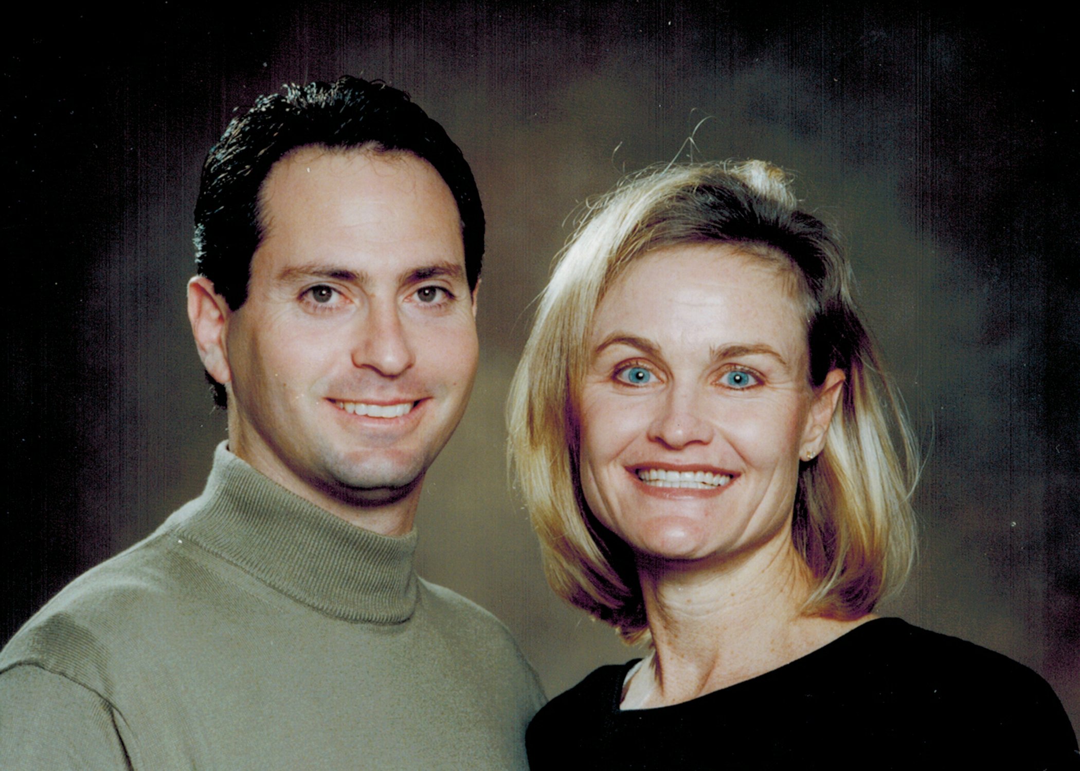 Close-up of a man with dark, slicked-back hair and a woman with blonde, shoulder-length hair and blue eyes, smiling together.