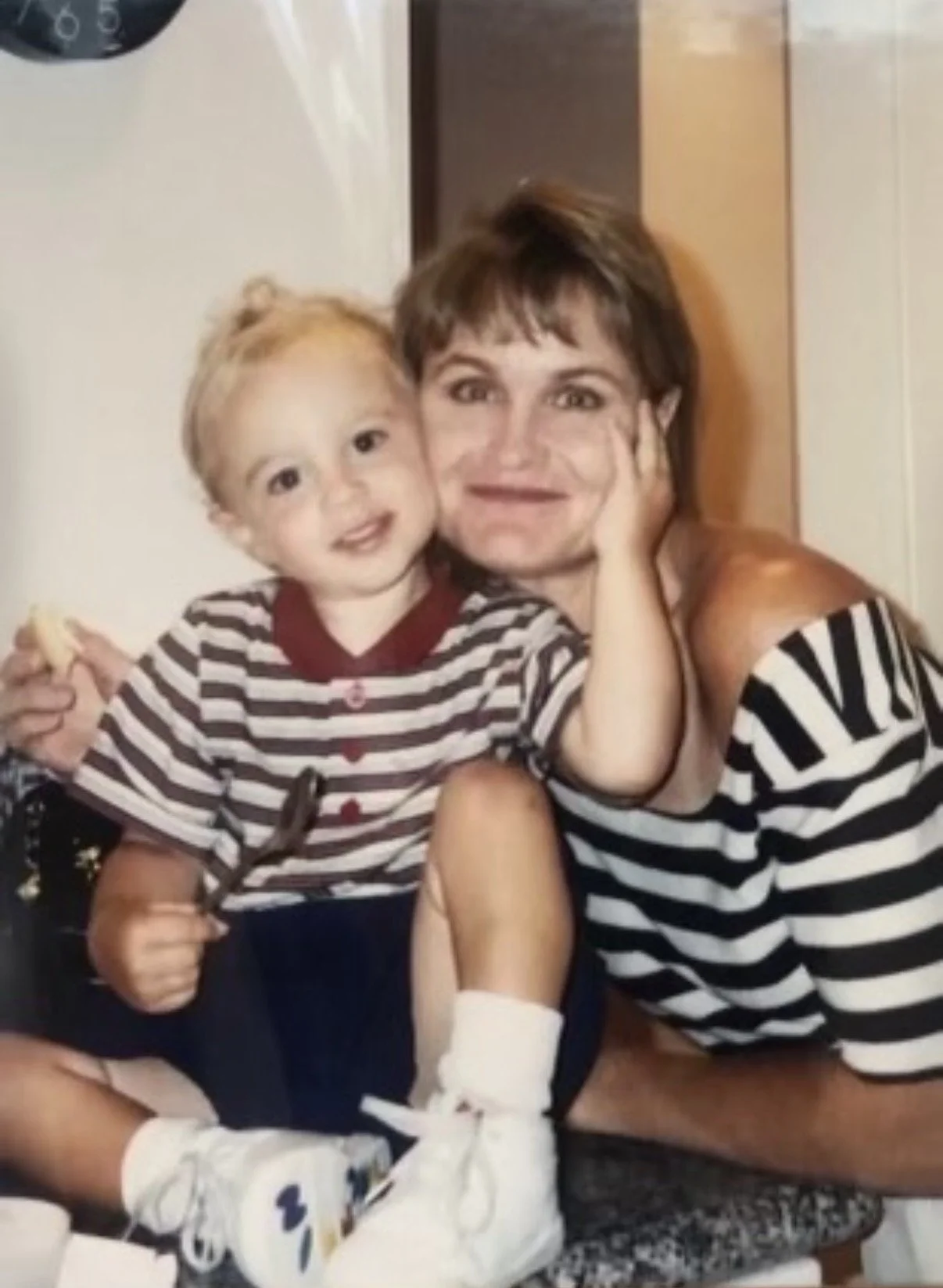 A woman and a young boy sitting on a stool, both smiling. The woman has short brown hair and is wearing a black-and-white striped dress. The boy has blonde hair, is wearing a three-quarter sleeve striped shirt, dark pants, and white sneakers. The wom