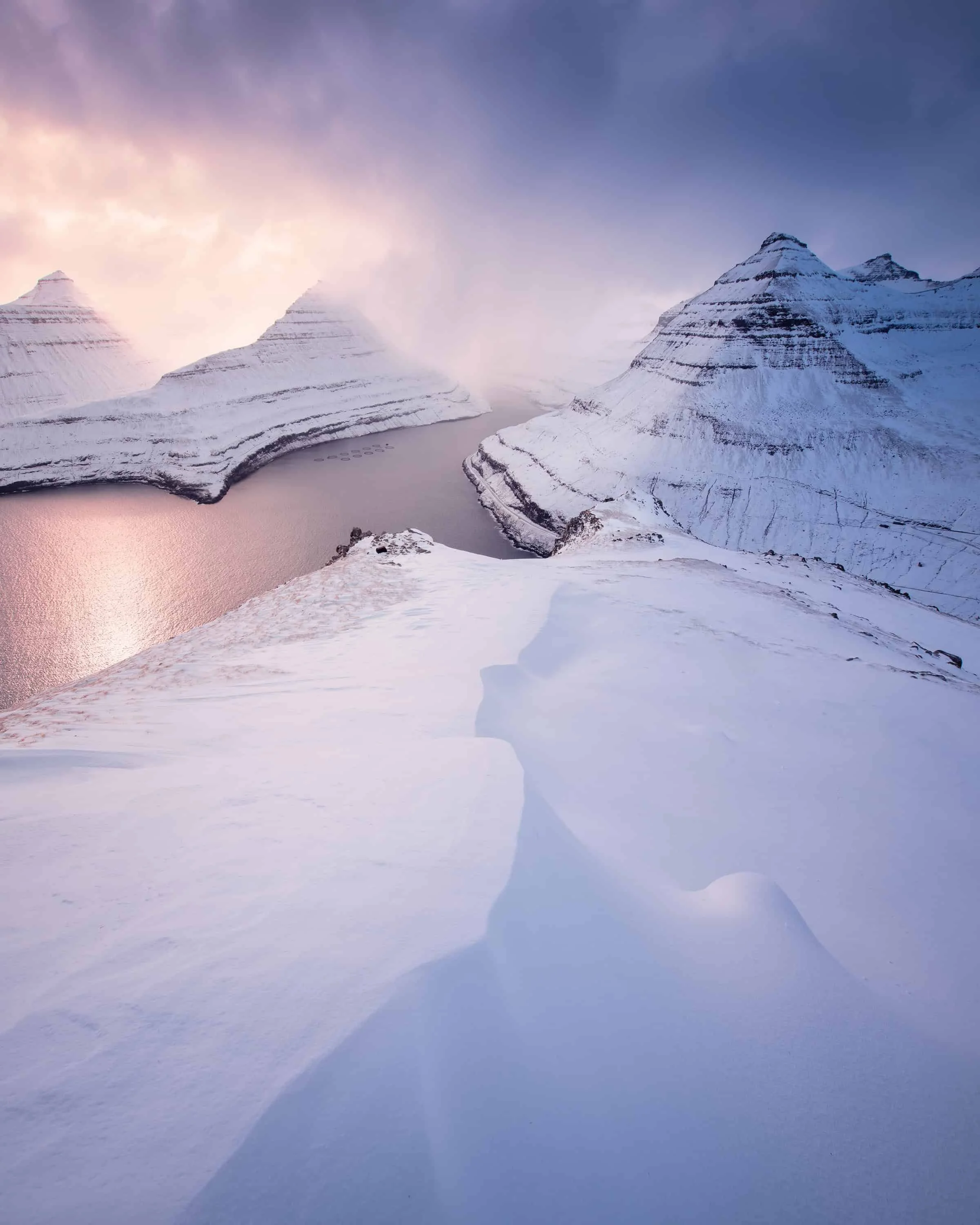 We’re overlooking Funningursfjørður on Eysturoy, Faroe Islands, in a rare moment of heavier-than-usual snowfall. Smooth snowdrifts in the foreground lead into the fjord, while the surrounding peaks stack in gentle layers under pastel sunset tones. Th