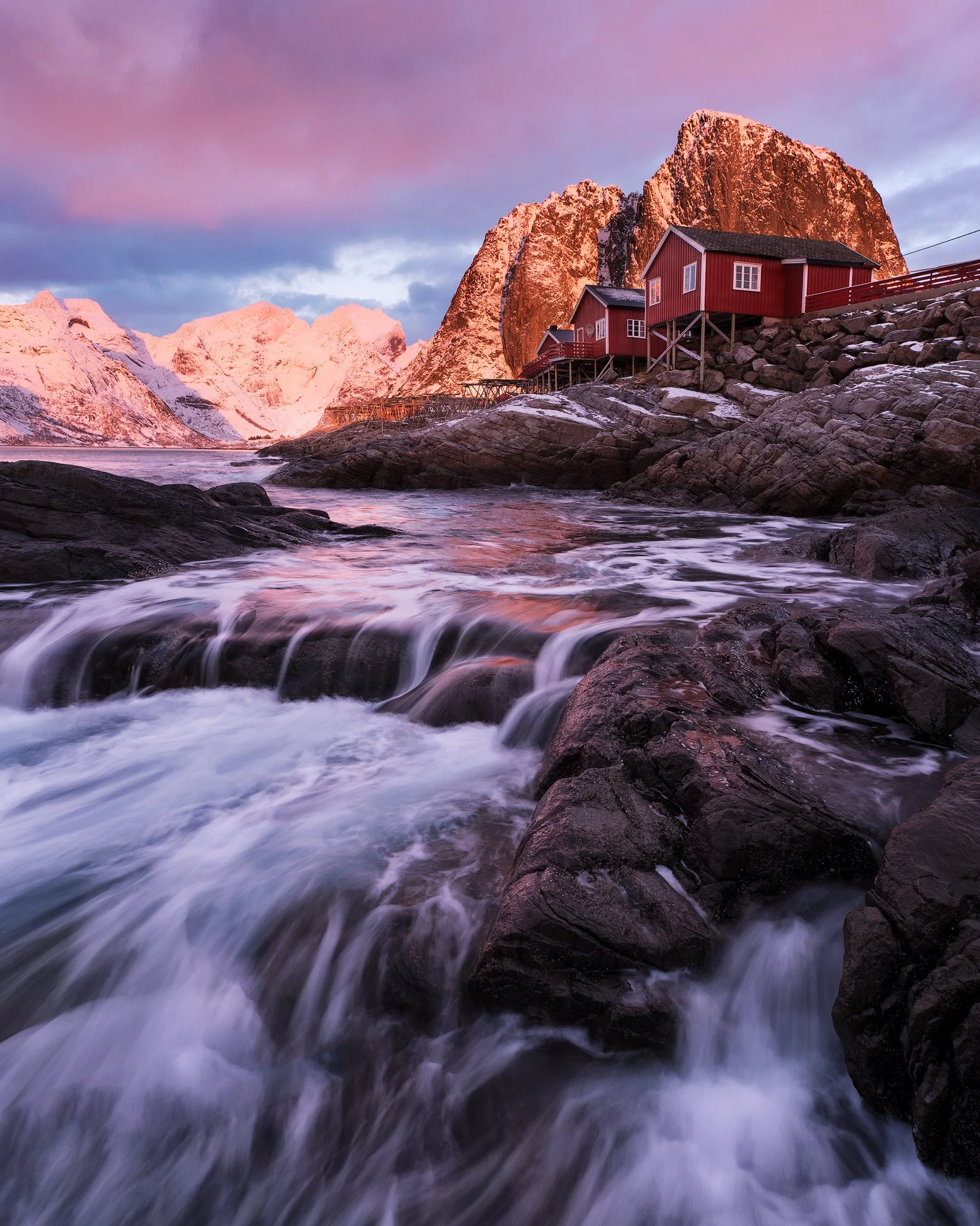Red rorbu below alpenglow peaks in Lofoten with water flowing over rocks at dusk