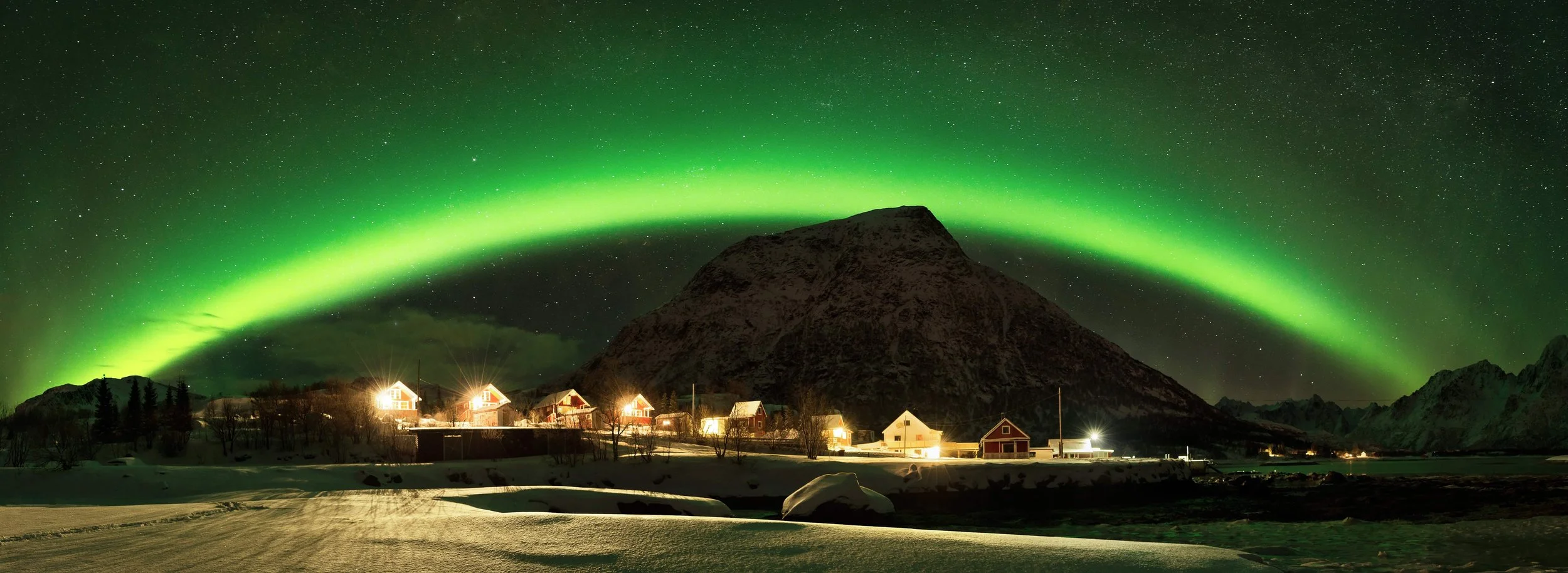 A tiny village in northern Norway under an arch of the northern lights captured by Felix Inden in a stitched panorama.