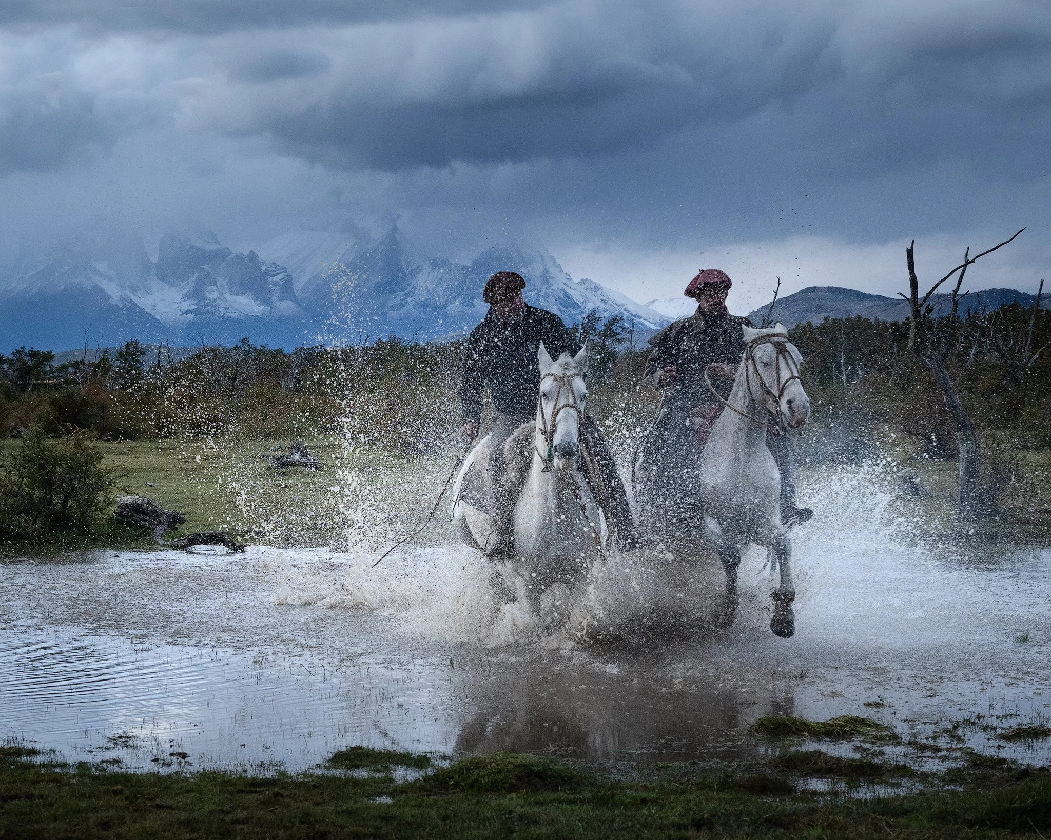 Two gauchose galopping through shallow water in front of the Cuernos del Paine in Torres del Paine National Parc in southern Chile