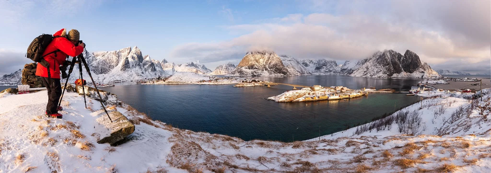 Photographer at a tripod on a snowy ridge overlooking Reine and Sakrisøy in winter, golden morning light on the snow-covered peaks, Lofoten, Norway