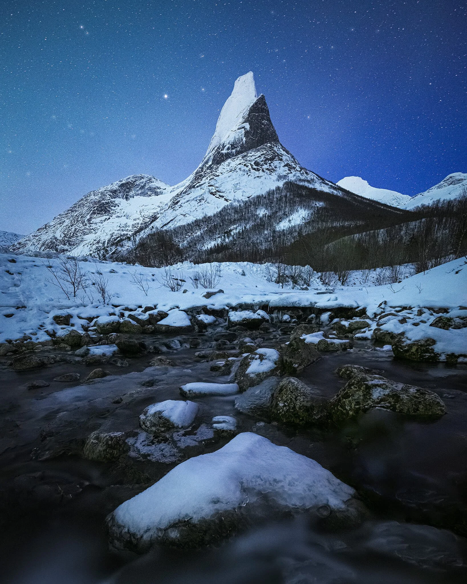 The sharp snow-covered spire of Stetinden under a starry night sky, with an icy, rock-strewn stream running through the snowy foreground.