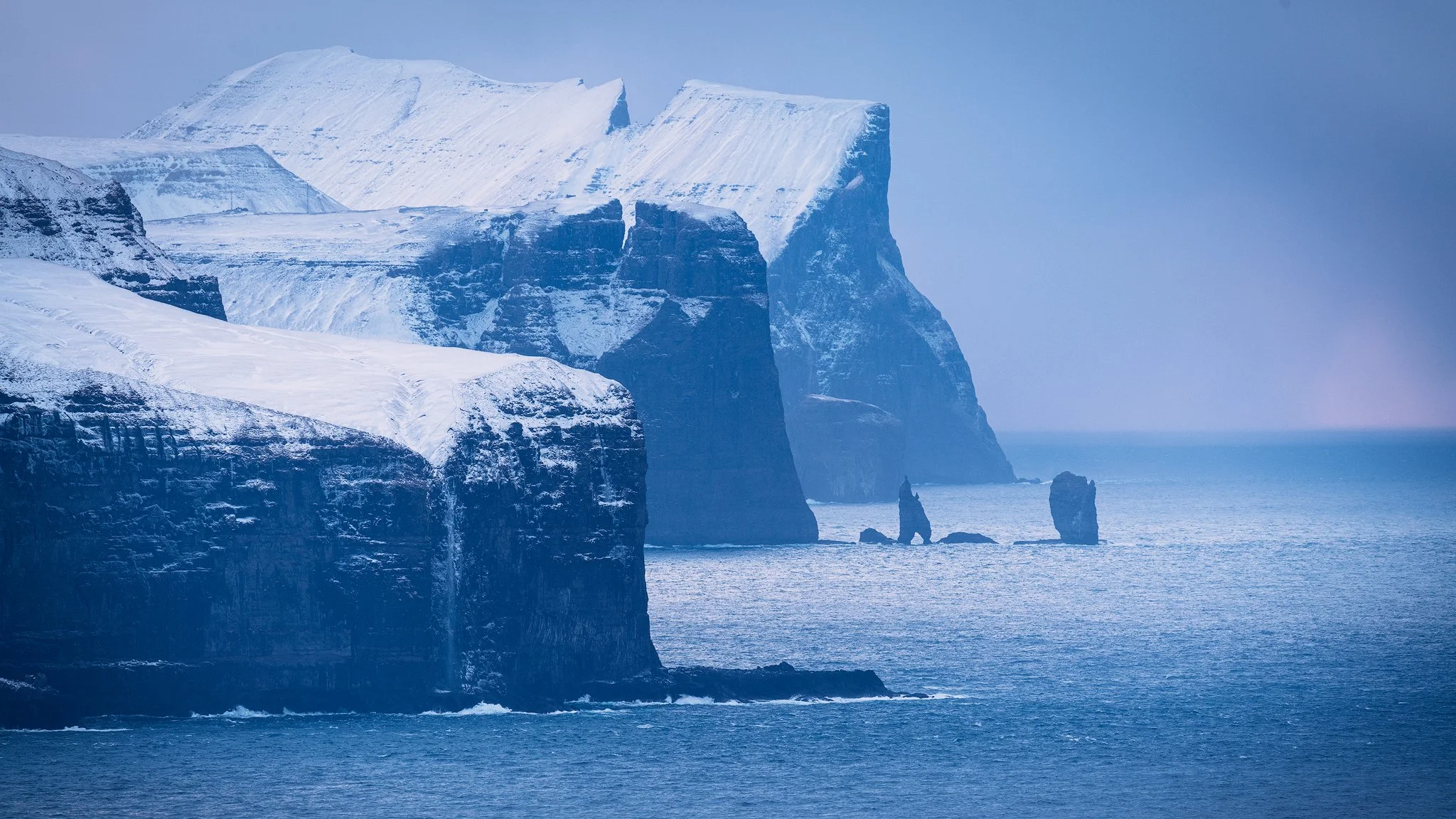 Telephoto winter view from Kalsoy toward snow-capped cliffs and the Risin og Kellingin sea stacks, Faroe Islands