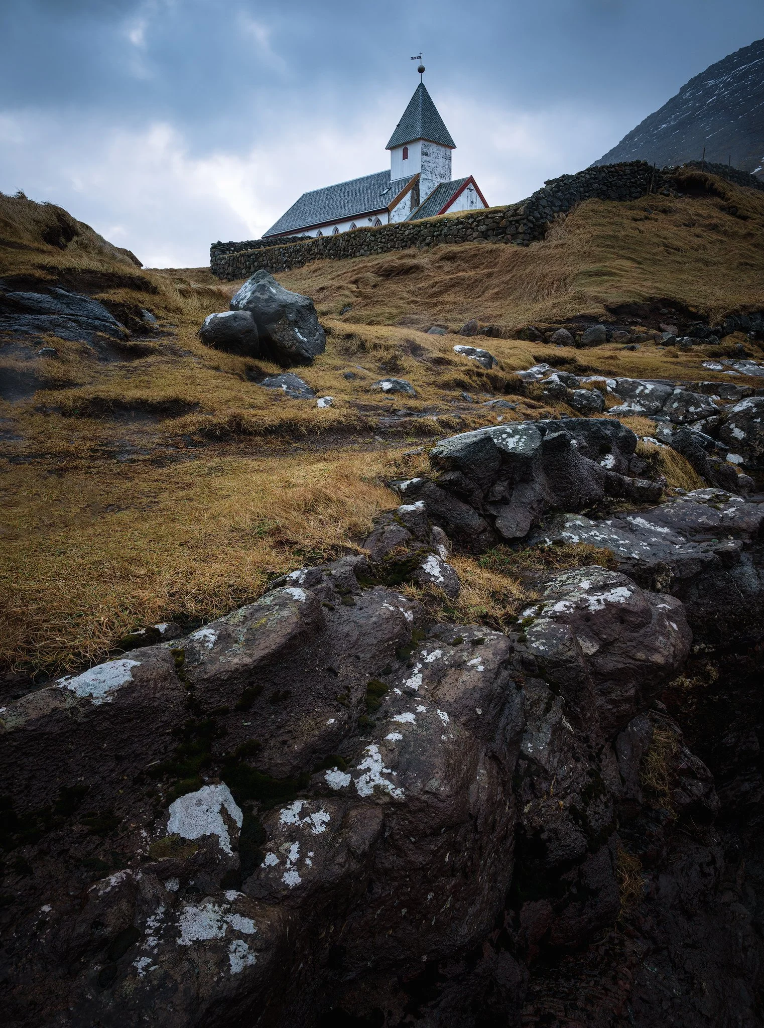 Low-angle view of the white church at Viðareiði with foreground rocks under a moody sky, Faroe Islands