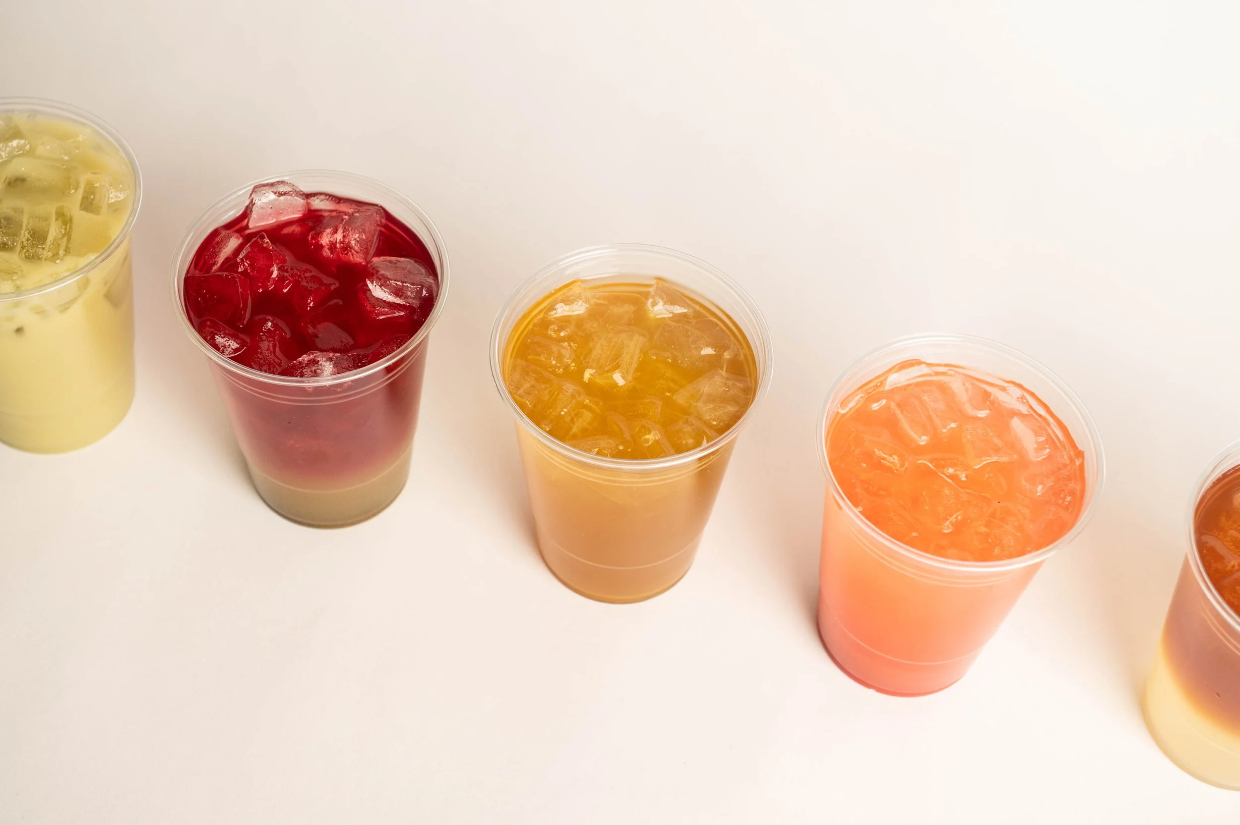 Four cups of colorful iced beverages on a white surface, with varying shades of yellow, red, orange, and pink.