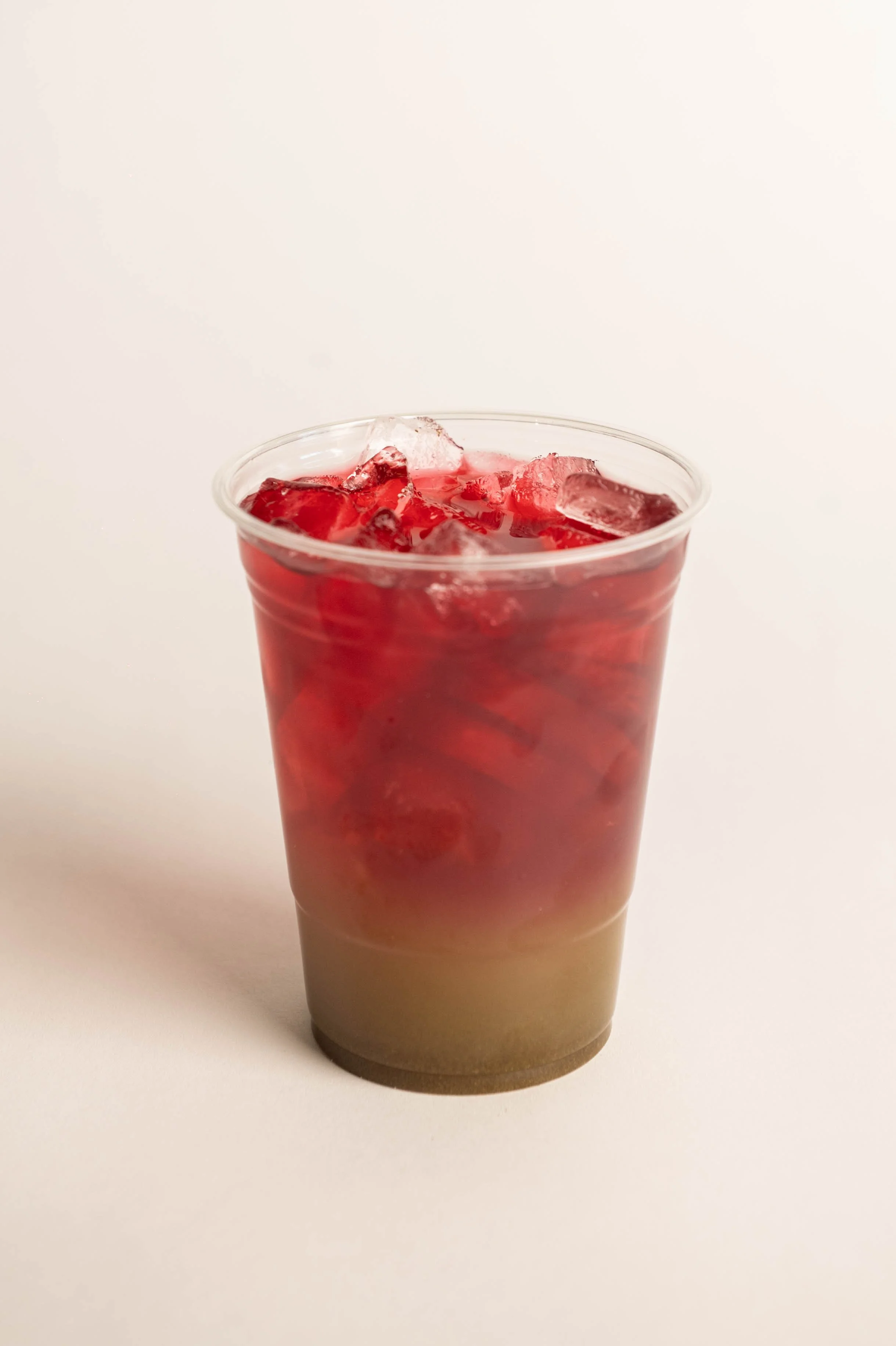 A clear plastic cup filled with a red beverage and ice cubes, placed on a white surface with a plain white background.