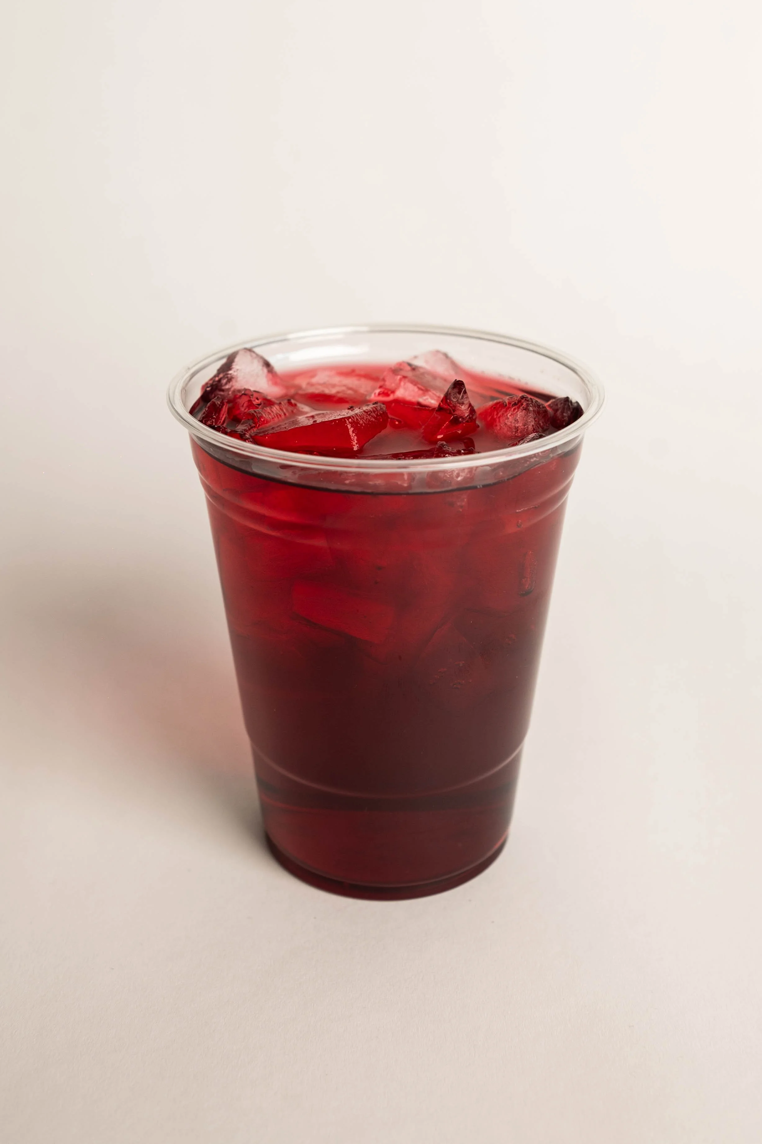 A clear plastic cup filled with red-colored beverage and ice cubes, placed on a white surface.