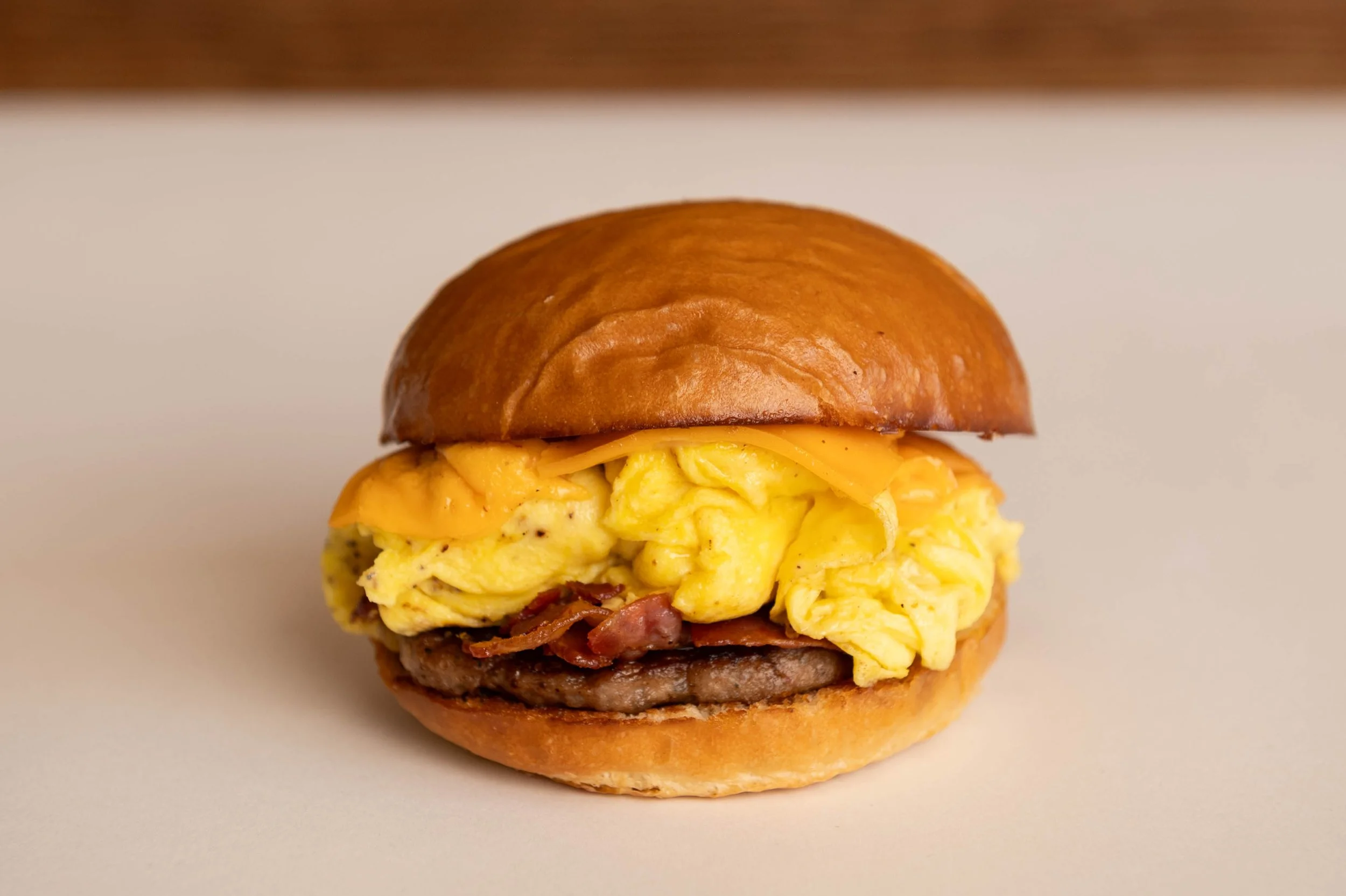 Close-up of a breakfast sandwich with scrambled eggs, cheese, bacon, sausage patty, in a toasted bun.