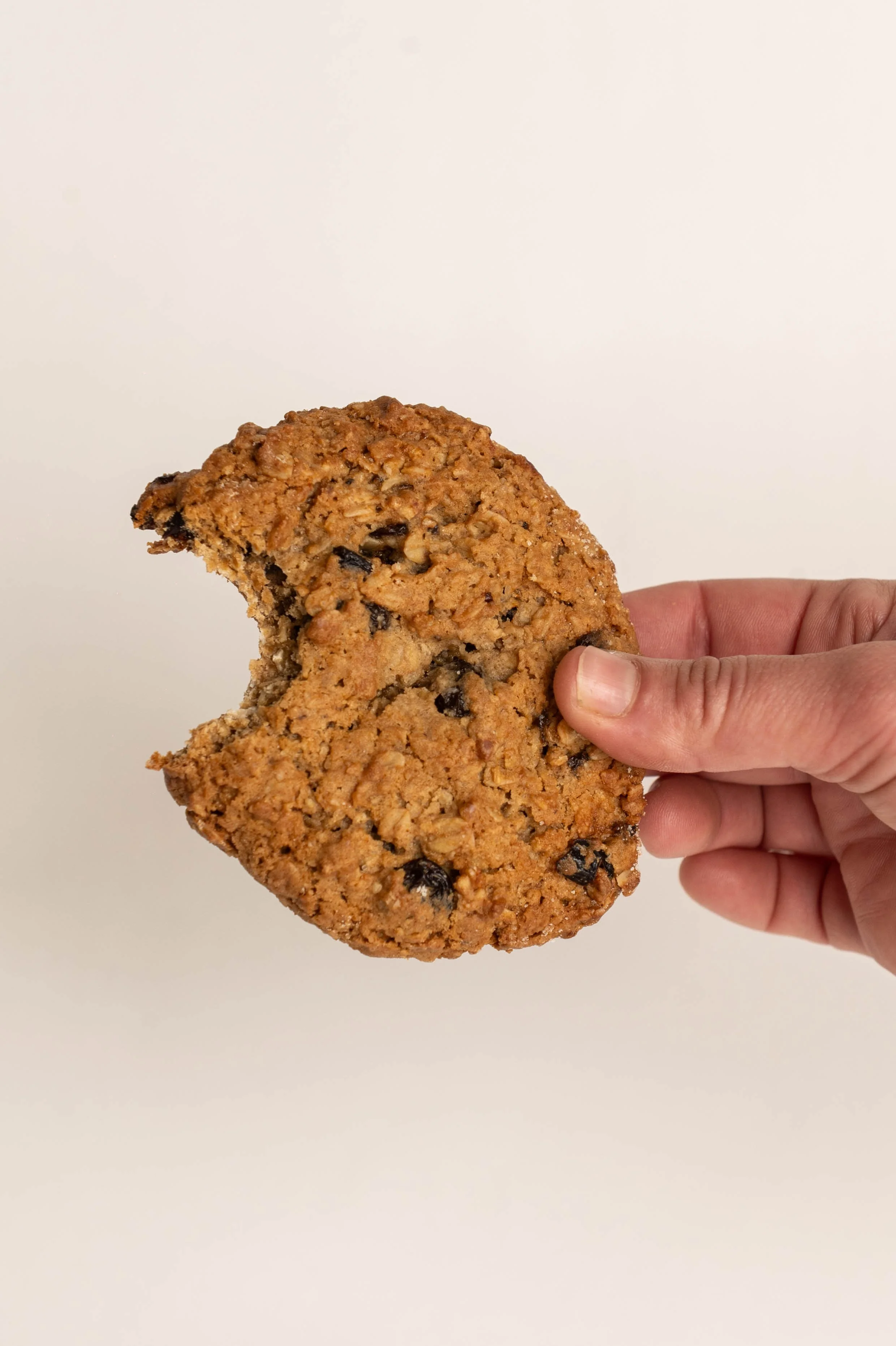 A hand holding a large oatmeal raisin cookie with a bite taken out of it, against a plain light background.