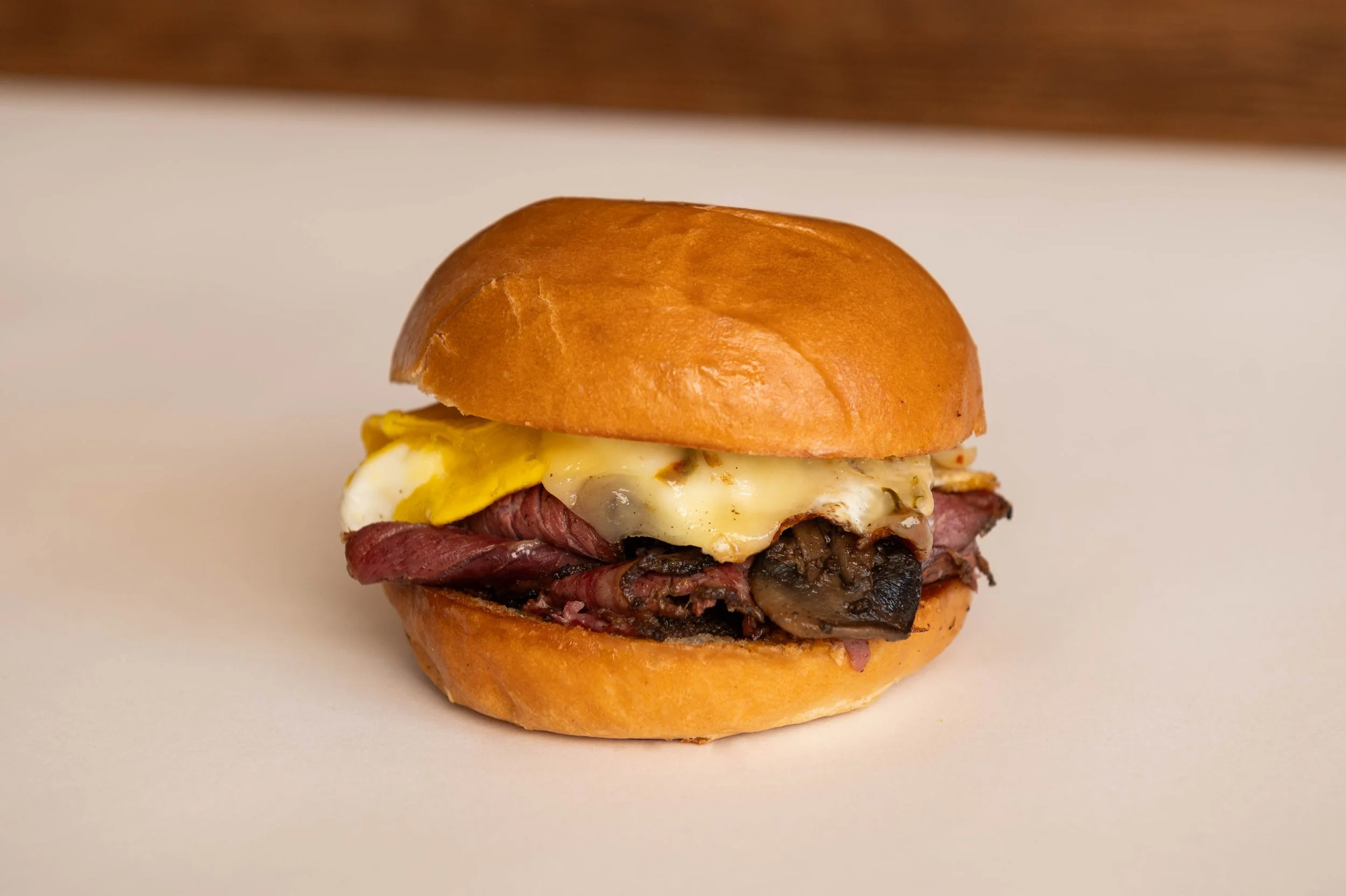 Sandwich with a toasted bun, fried egg, roast beef, and mushrooms on a white surface.