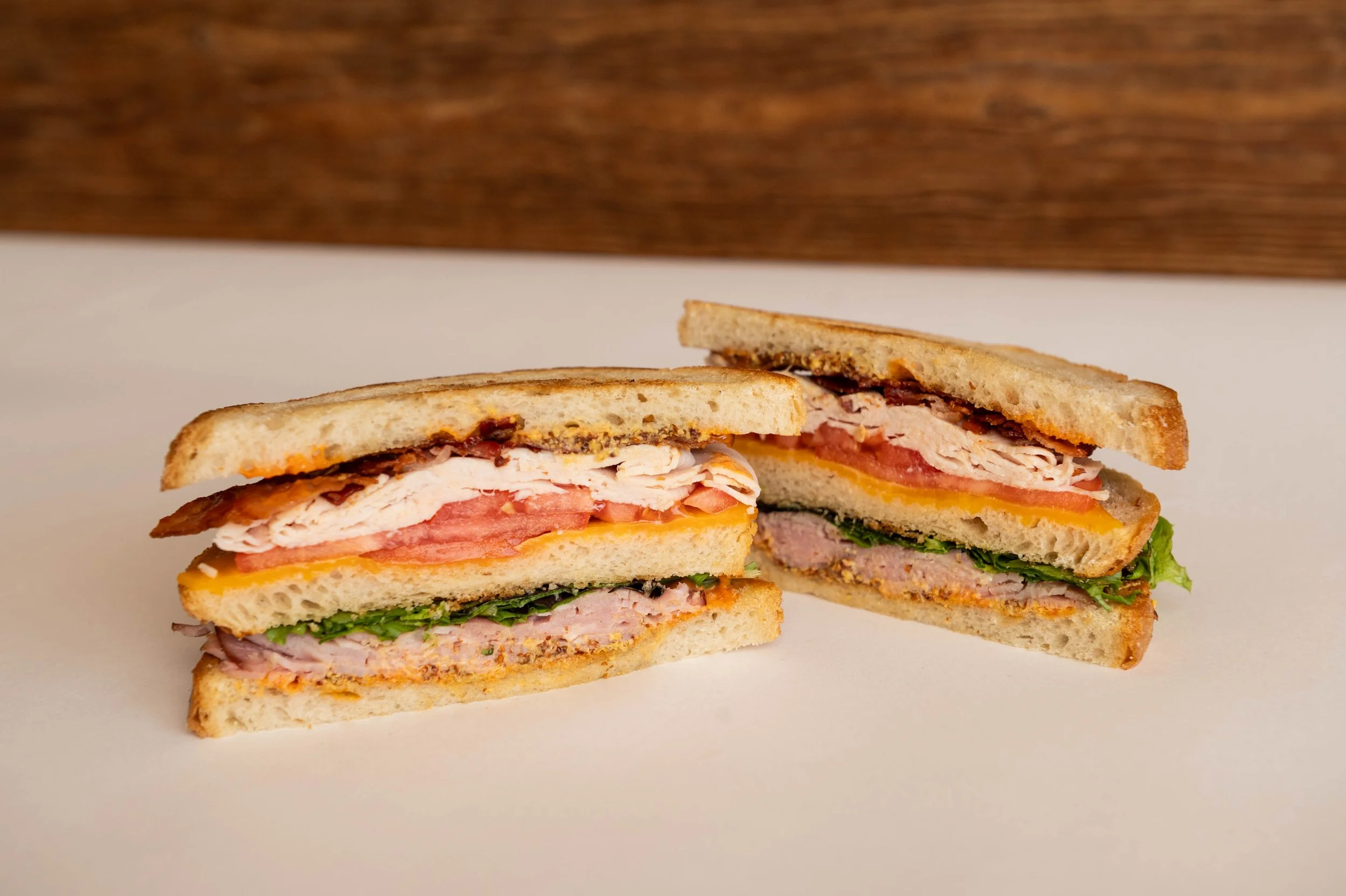 Half and quarter of a sandwich with turkey, ham, bacon, cheese, lettuce, tomato, and bread on a white surface with a wooden background.