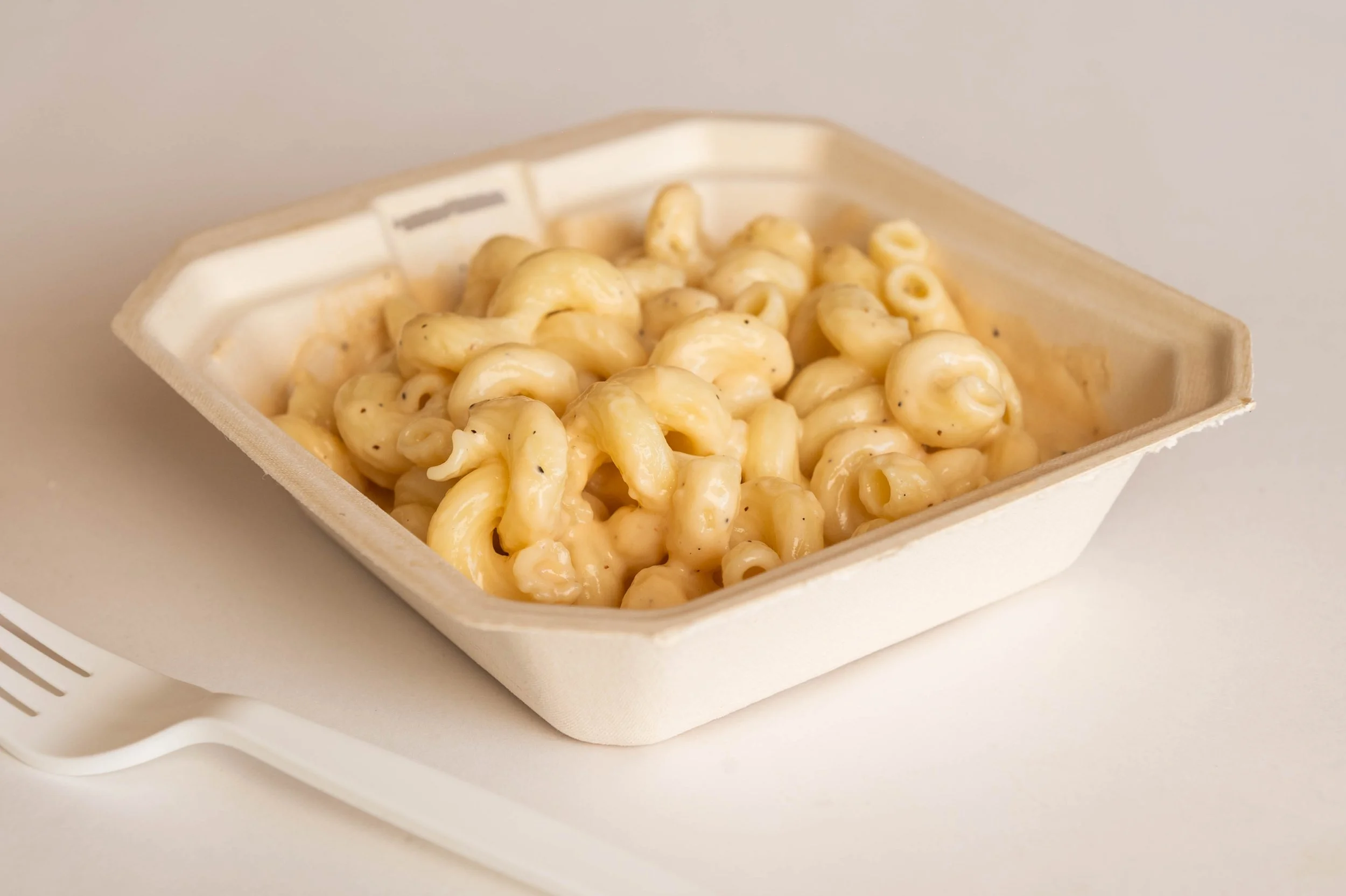 To-go container filled with macaroni and cheese on a white surface, with a white fork beside it.