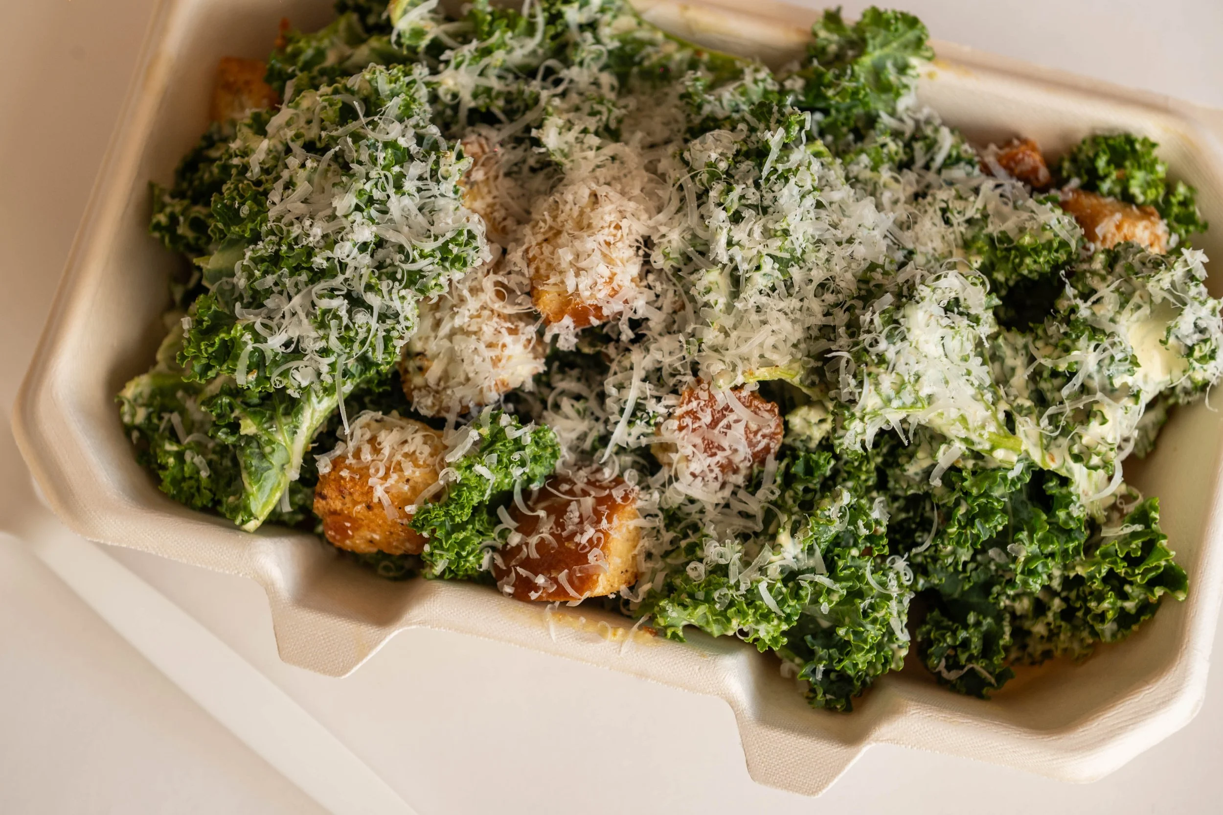 Kale Caesar salad with shredded cheese and croutons in a takeout container.