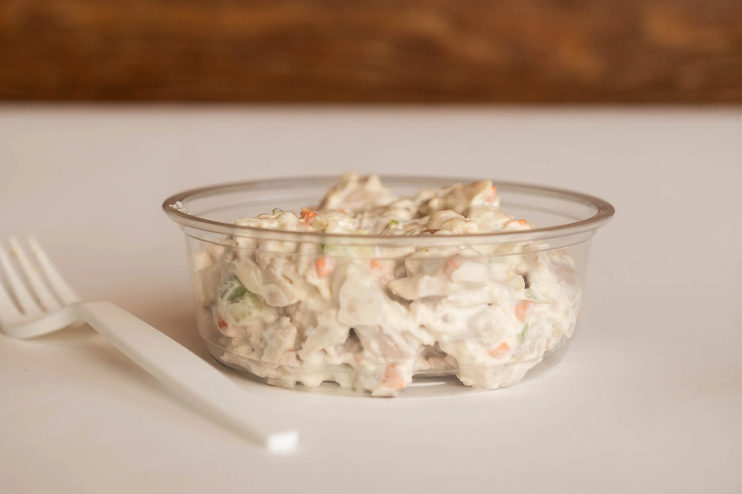 A clear plastic container filled with creamy chicken salad with visible pieces of chicken, celery, carrots, and mayonnaise, placed on a white surface next to a white plastic fork.
