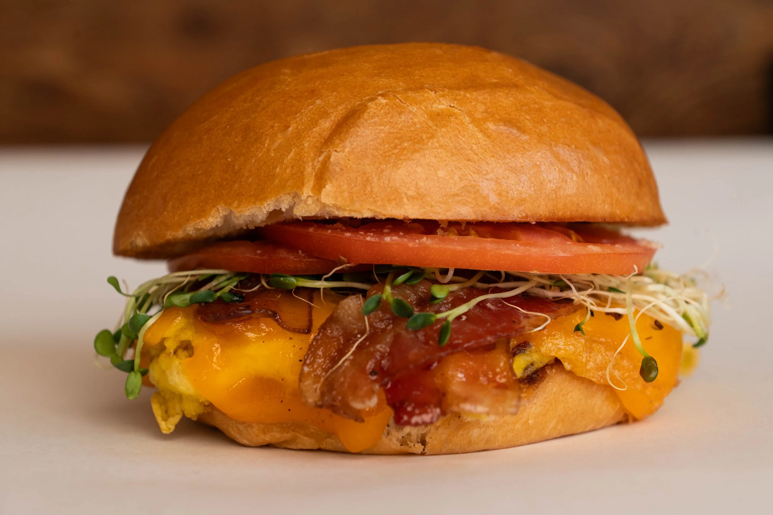 Close-up of a breakfast sandwich with egg, cheese, bacon, tomato, and sprouts on a bun.