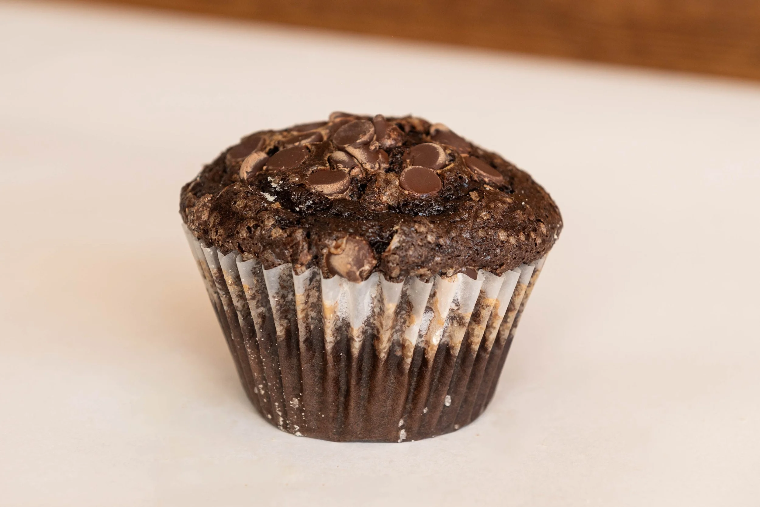 Chocolate muffin with chocolate chips on top, in a white paper cup, placed on a white surface.