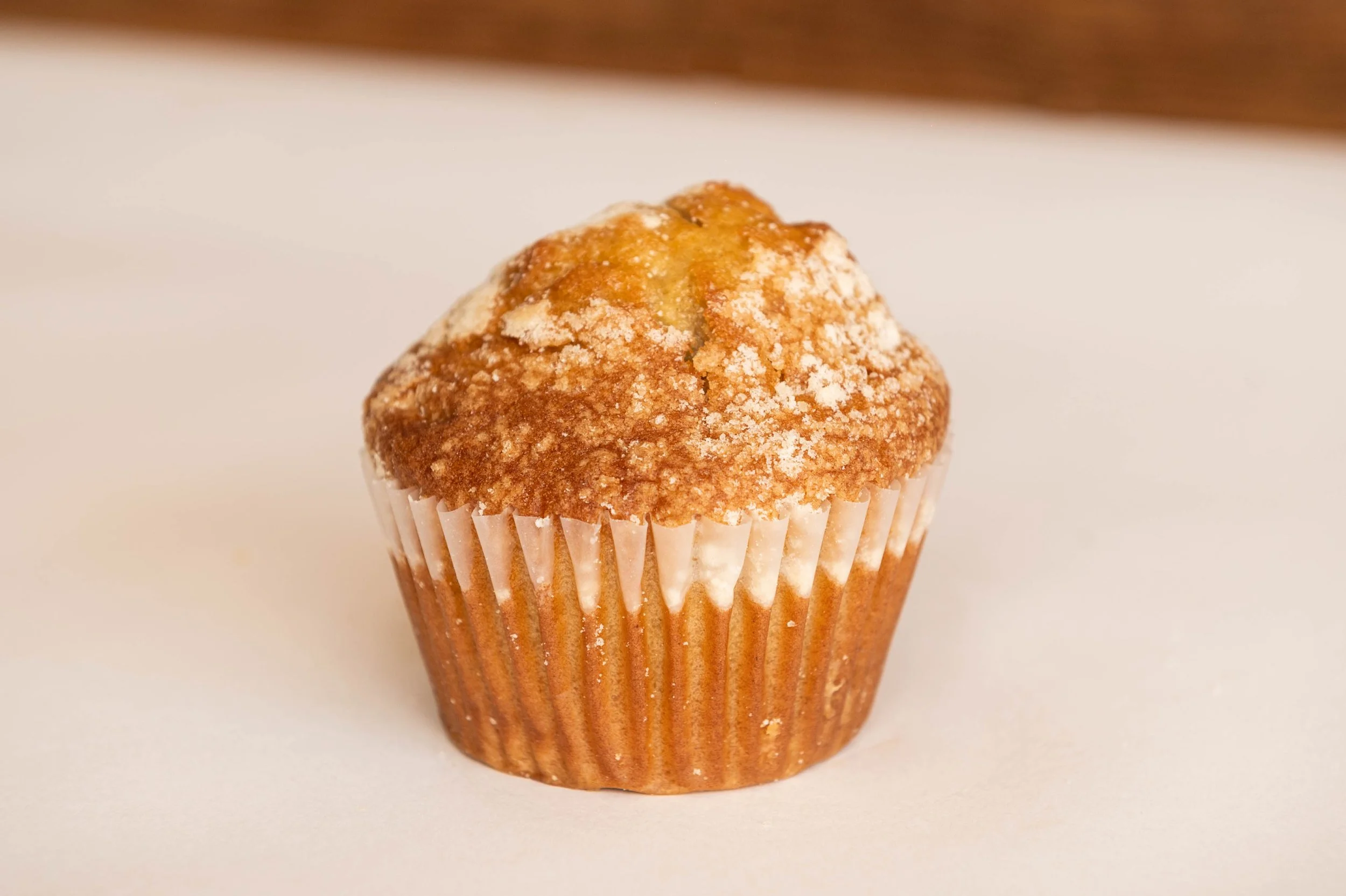 A close-up of a single muffin with a golden-brown top dusted with powdered sugar, placed in a white paper liner on a white surface.