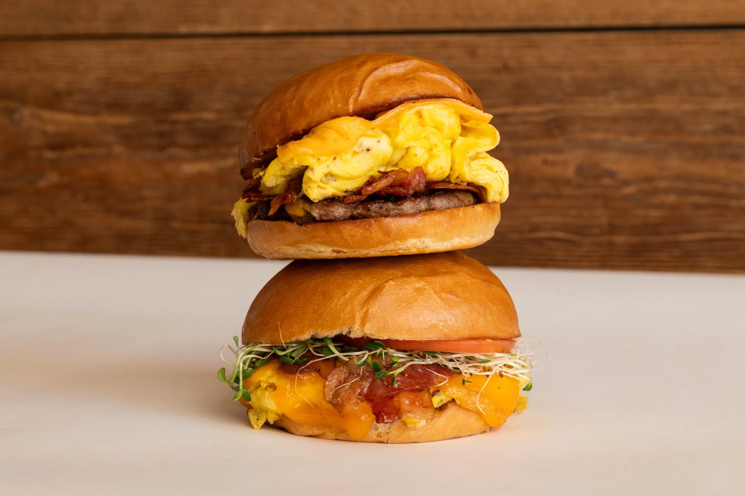 Two stacked breakfast sandwiches with scrambled eggs, bacon, cheese, tomato, and microgreens on sandwich buns against a wooden background.