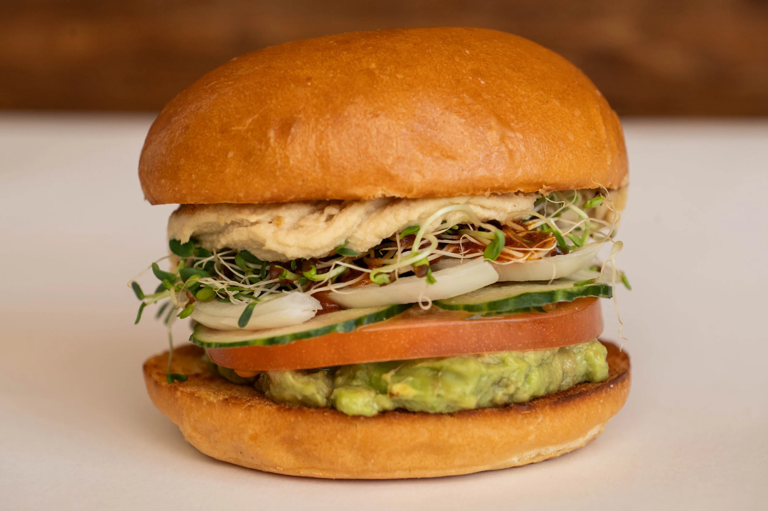 Close-up of a sandwich with a toasted bun, guacamole, tomato, cucumber, sprouts, sliced onion, and a fried egg.