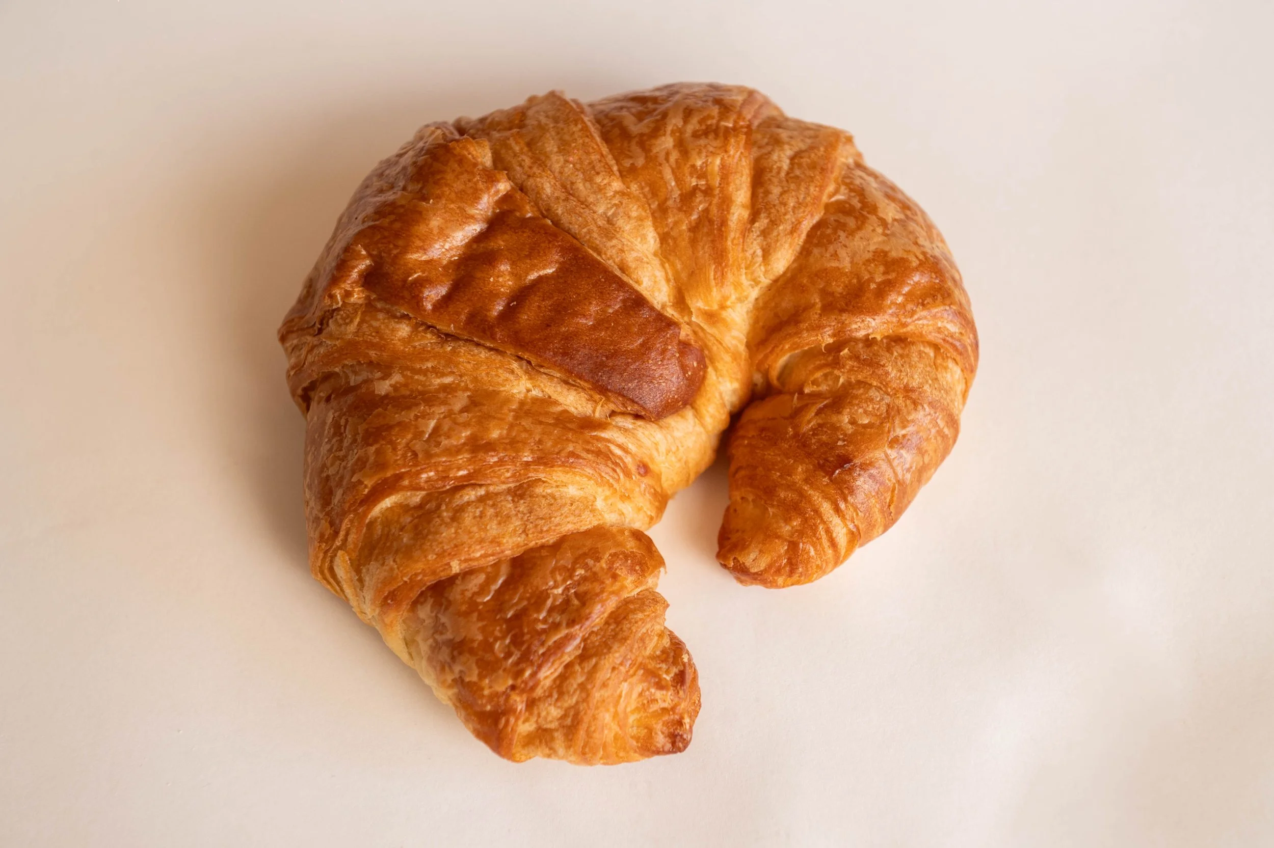 A freshly baked croissant with a slightly darker baked triangle topping, placed on a plain, light-colored surface.
