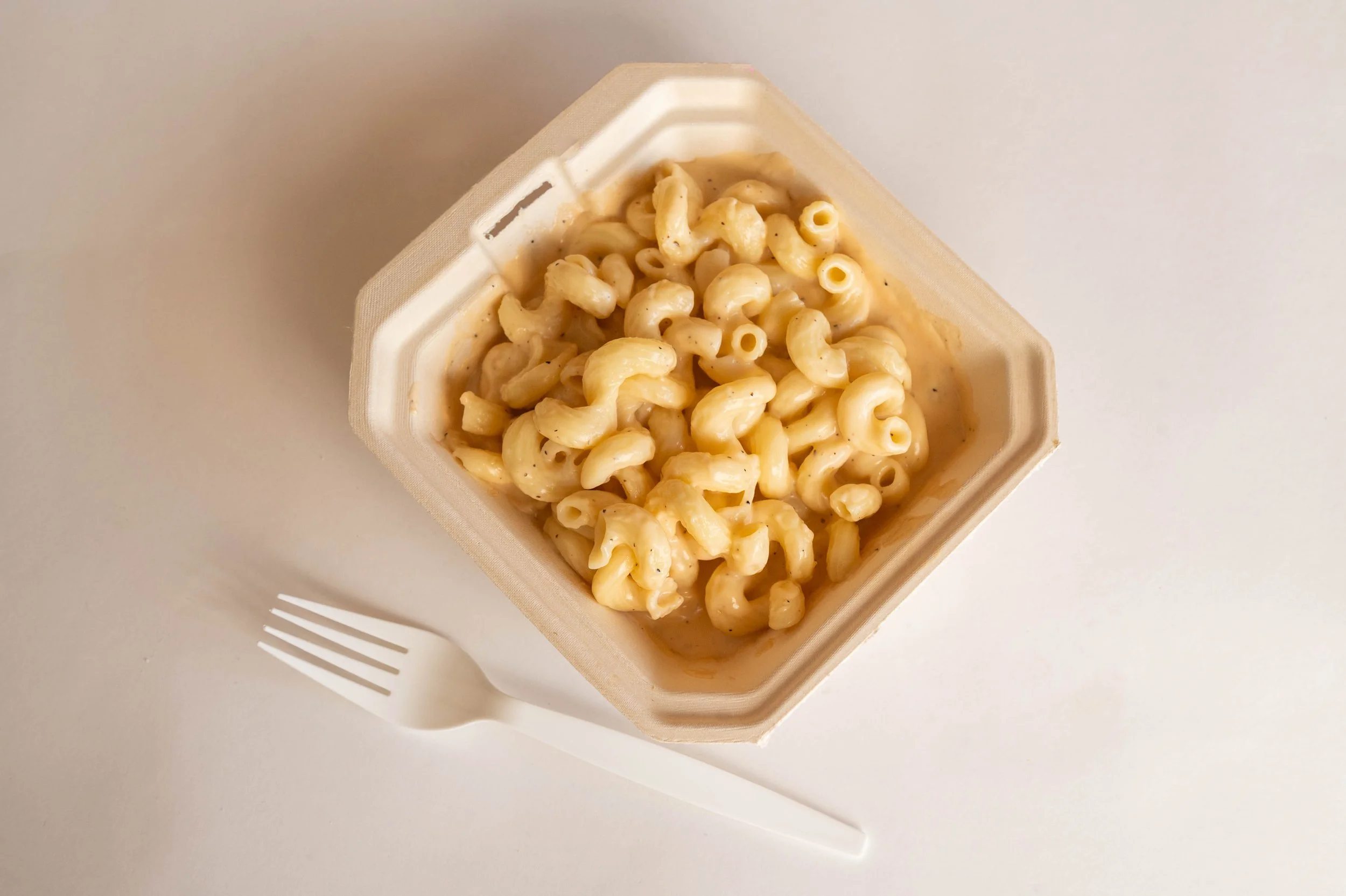 A white foam container filled with macaroni and cheese on a white table, with a white plastic fork beside it.