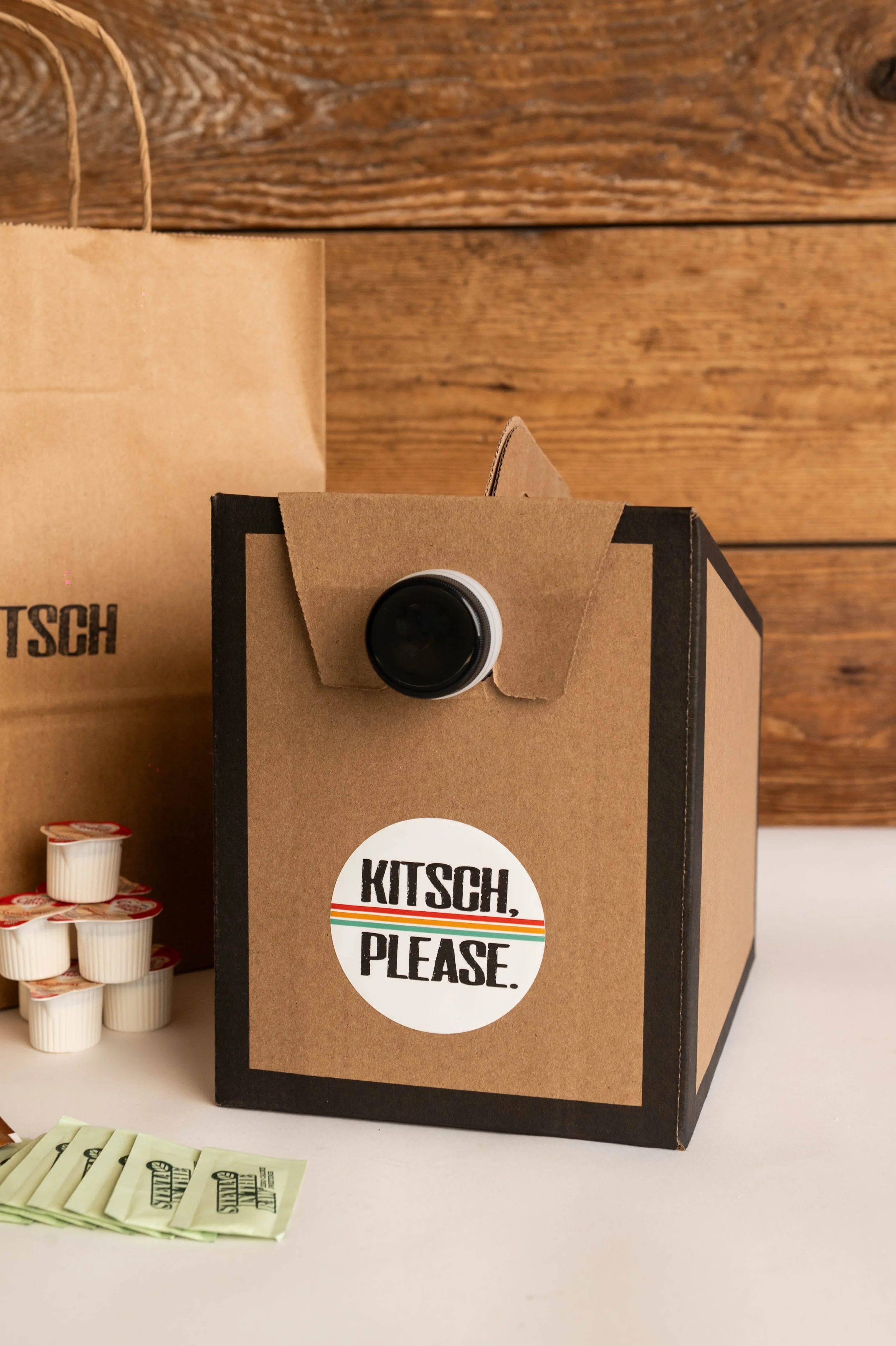 Coffee sampling box with a camera lens on the front, logo reading "Kitsch, Please," pods, napkins, and a paper bag on a counter against a wooden background.