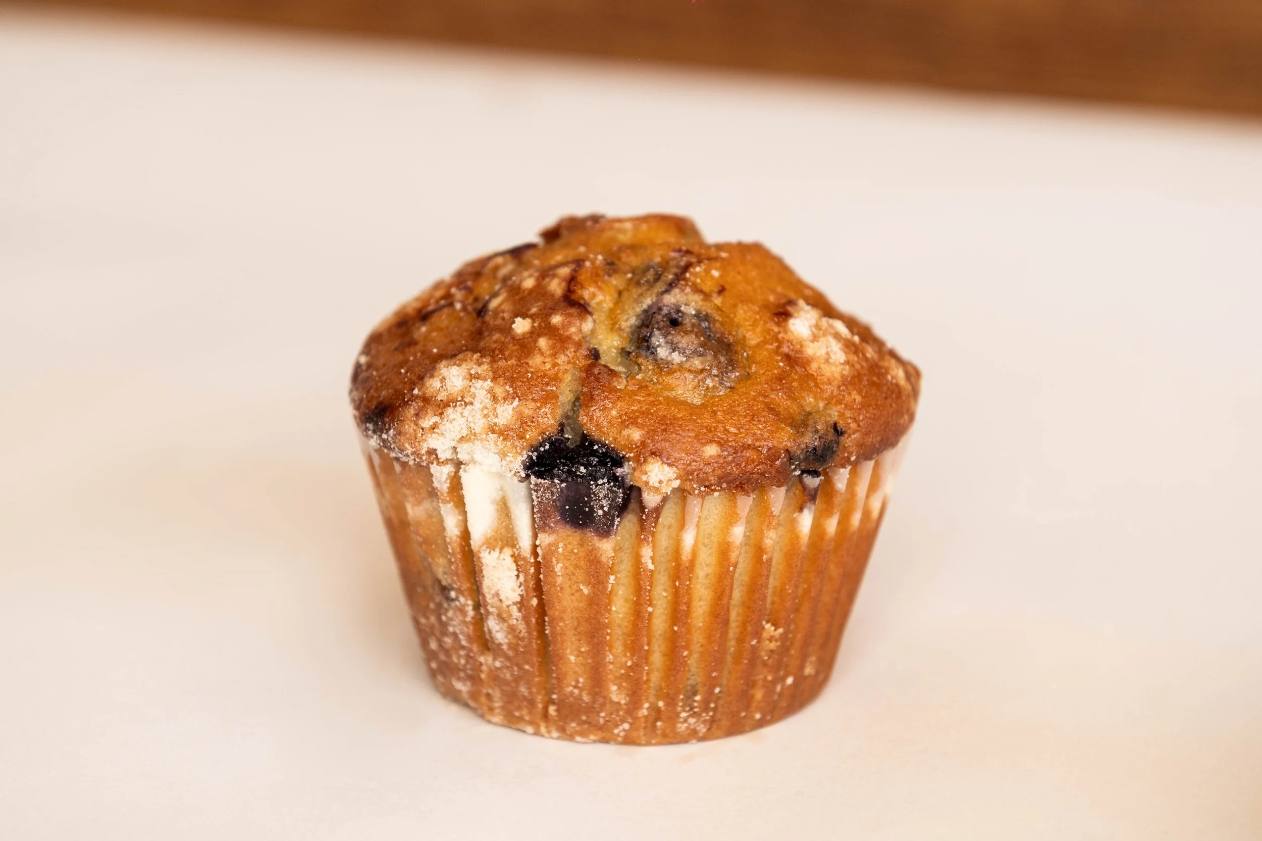 A blueberry muffin with a golden-brown top, slightly cracked, dusted with powdered sugar, on a white surface.