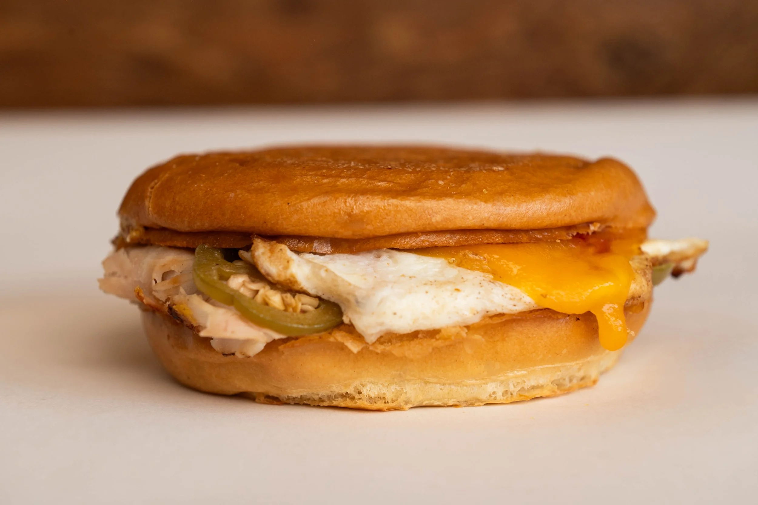 Close-up of a breakfast sandwich with fried egg, cheese, sliced pickles, and meat on a toasted bun.