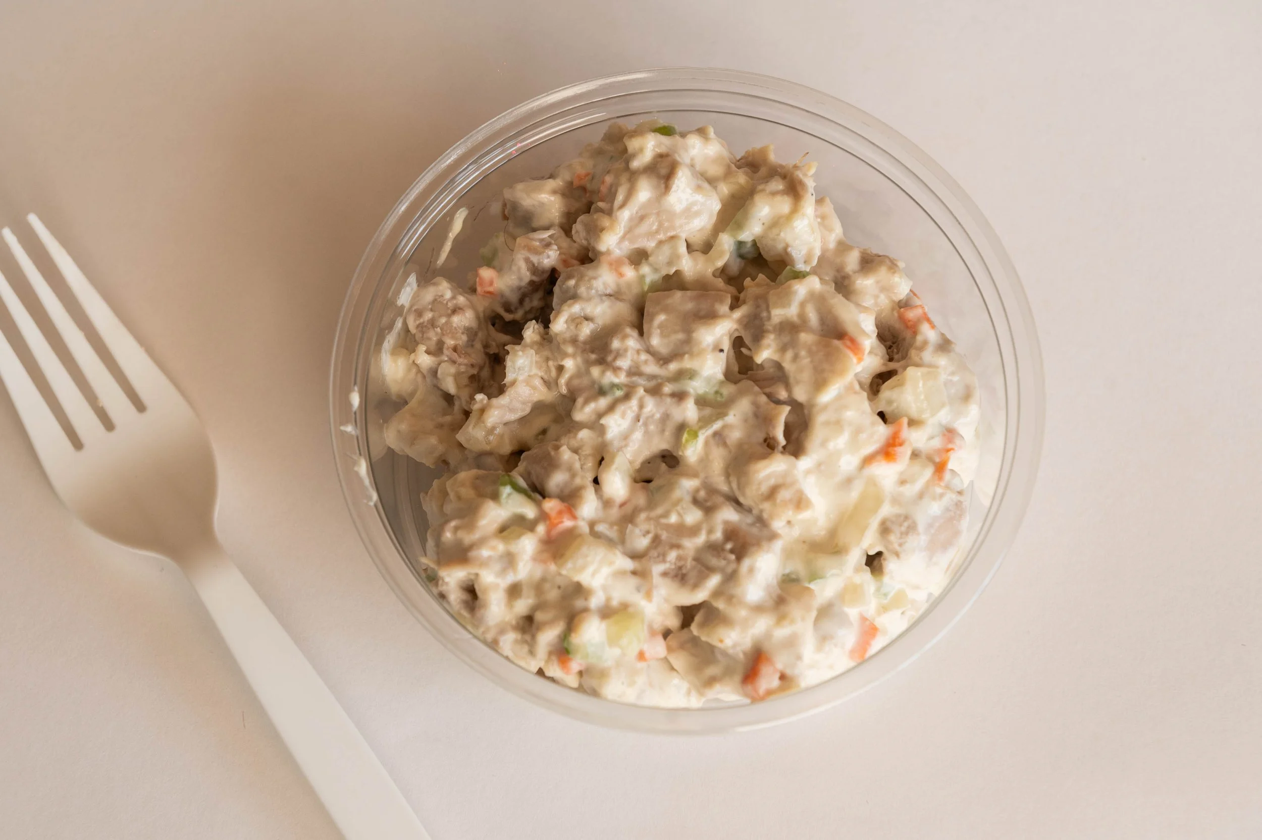 A bowl of creamy chicken salad with chopped vegetables on a white surface, with a white plastic fork beside it.