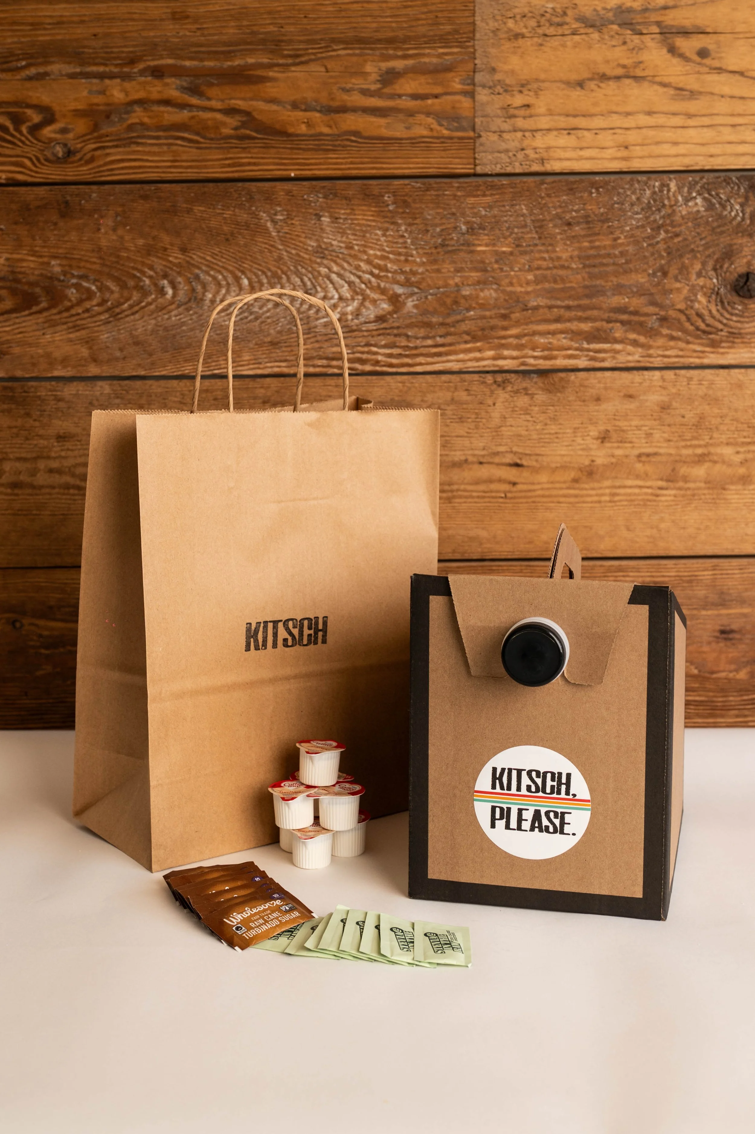 A paper bag, a small cardboard box with a camera lens on top and a label saying "KITSCH, PLEASE," along with individual cream container packets and sugar packets on a white surface with a wooden wall background.